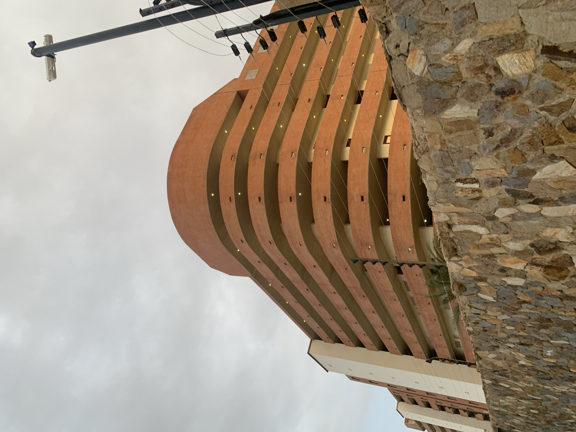 The image displays a multi-story building featuring a distinctive, curved architectural design under an overcast sky. The building, constructed from a reddish-brown, brick-like material, showcases multiple layered, sweeping balconies or facade elements that recede inwards. Small, warm-toned lights are visible embedded along the curves of these structures.
A rugged wall or pillar made of varied-sized natural stones in earthy tones occupies the far right side of the frame, partially obscuring the building. In the upper left quadrant, a dark utility pole extends into the shot, supporting electrical wires and a small, white rectangular device, possibly a security camera or wireless antenna.
The background consists of a uniformly light grey, cloudy sky, indicating an overcast day. No individuals or specific activities are visible within the frame, emphasizing the architectural details and the urban or resort-like setting. The scene is located in Pampatar, Venezuela. There is no discernible text in the image.