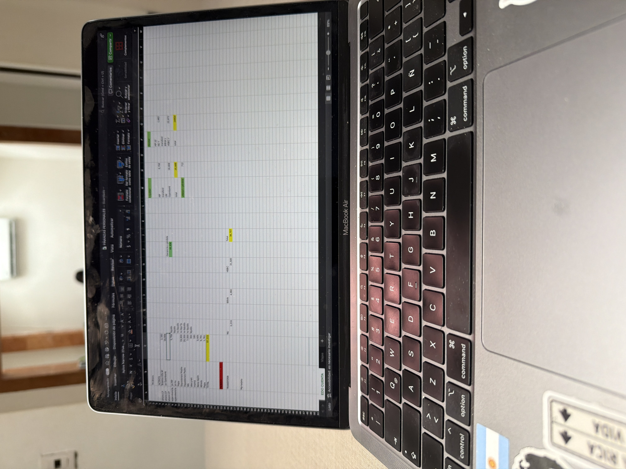 This is a close-up, vertical shot of a MacBook Air laptop open, displaying a spreadsheet on its screen. The laptop is positioned on a surface that appears to be a desk or table. The scene is indoors, and the lighting suggests it could be daytime.
The spreadsheet on the screen contains financial data, with columns and rows filled with numbers and text, primarily in Spanish. Visible text includes headings like "FINANZAS PERSONALES" (Personal Finances), "ACTIVOS" (Assets), and financial institutions such as "BBVA" and "HSBC." There are also numerical values and categories like "Ganancia o pérdida" (Profit or loss).
The keyboard of the MacBook Air is also prominently visible, with its standard layout. Two stickers are affixed to the bottom right of the laptop's casing. One is a small Argentinian flag, and another is a white sticker with some text, partially obscured but appearing to say "VIDA RICA".
The laptop is the central object of focus. No people are visible in the frame. The background is blurred, showing hints of a wall and possibly a window with natural light coming through, suggesting a domestic or office setting. The overall impression is one of someone working on personal finances. The cropped nature of the image emphasizes the laptop and its screen content.