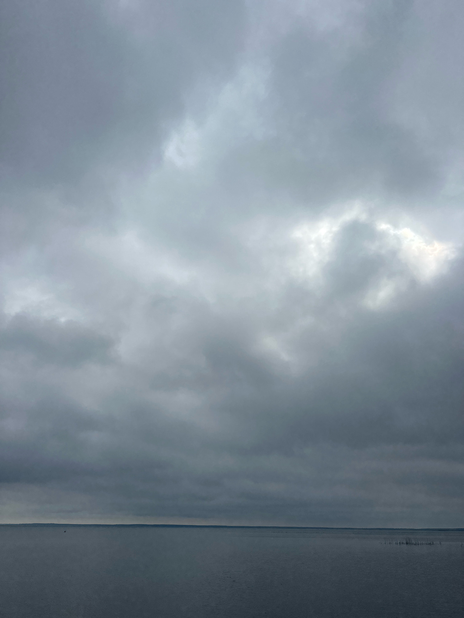 The image depicts a serene, overcast day over a body of water, likely a lake or a wide river. The sky is filled with dense, grey clouds, with a few brighter patches suggesting diffused sunlight. The water is calm and dark, reflecting the muted tones of the sky. A thin, dark line on the horizon indicates a distant shoreline. There are no visible people or significant man-made structures in the immediate vicinity of the water. The overall atmosphere is tranquil and somewhat somber due to the overcast conditions. The location is identified as Нарацкі сельскі Савет, Belarus.