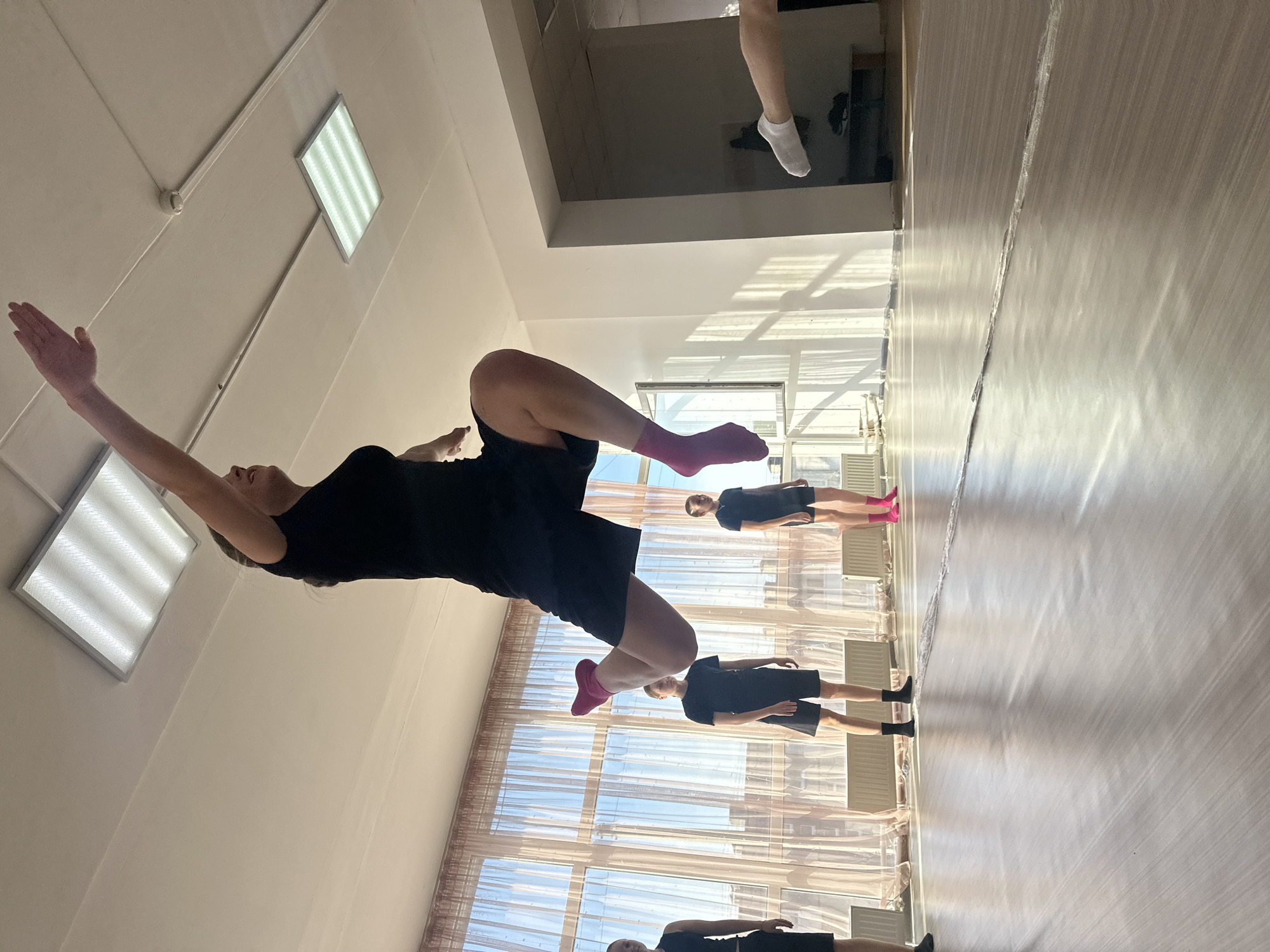 The image depicts a dance studio in Votkinsk, Russia. Several individuals, dressed in black outfits, are participating in a dance class. The foreground features a woman in mid-leap, her arm extended upwards. The background shows other dancers performing similar moves on the floor. The studio has large windows and fluorescent lighting on the ceiling. The floor is a light, polished wood.