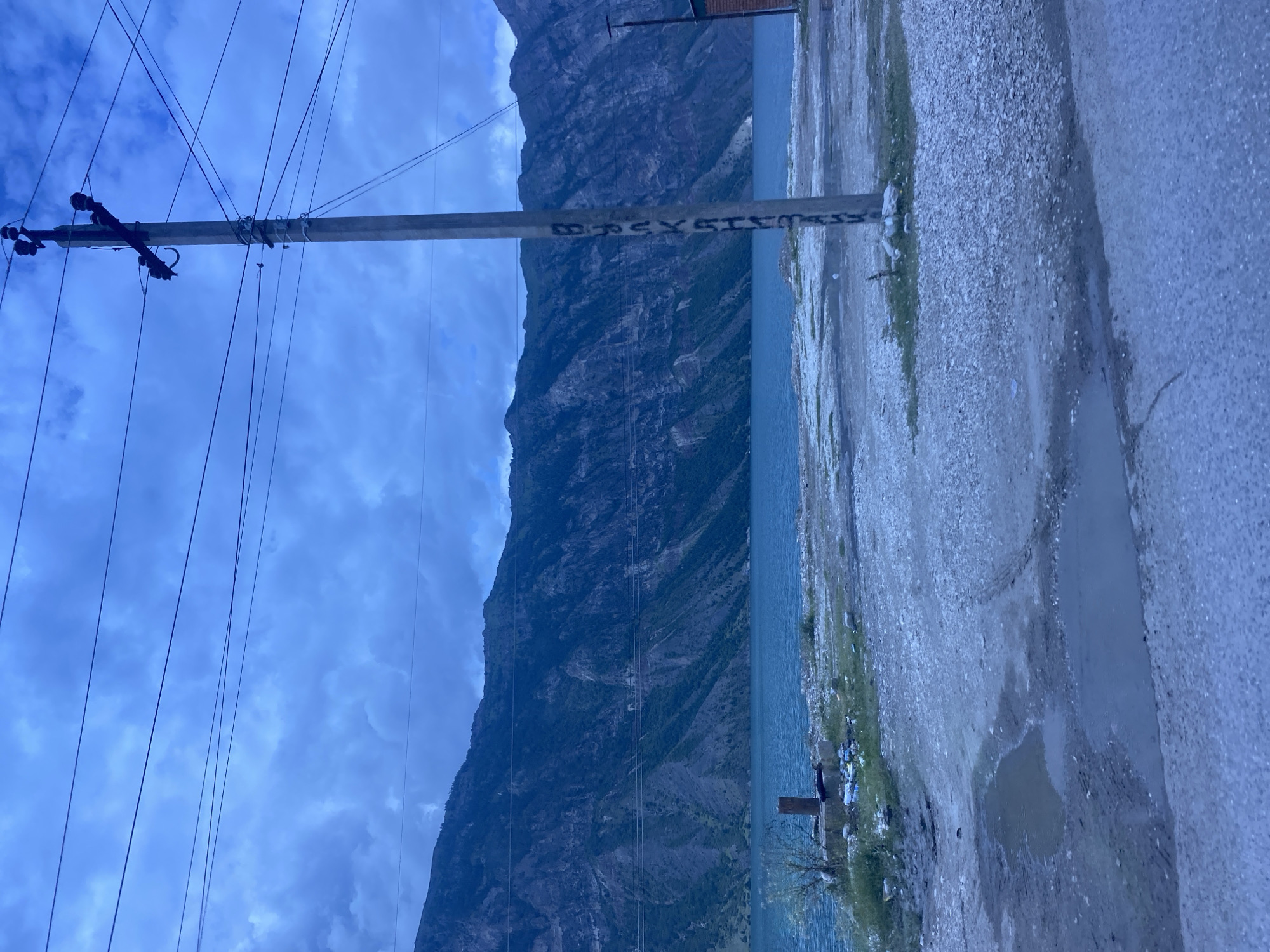The image depicts a dusky, overcast day in the vicinity of the Irganay reservoir in Irganay, Russia. A concrete utility pole with electrical insulators and wires is prominently featured in the foreground, with Cyrillic writing on its surface. The pole stands on a dirt and gravel embankment that slopes down to the bright blue waters of the reservoir. Beyond the reservoir, a massive, dark, and rugged mountain range dominates the background, its steep slopes etched with shadows and some sparse vegetation. The ground in the immediate foreground is littered with debris, including plastic bags and other refuse, and shows evidence of puddles and tire tracks. A small, unkempt patch of grass is visible near the base of the utility pole, with a low concrete structure and more scattered trash. To the far right, a portion of a brick building is visible. The overall impression is one of a somewhat neglected, natural landscape adjacent to man-made infrastructure.