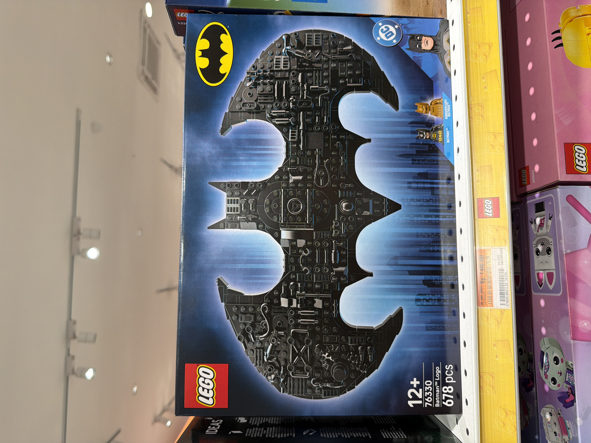 The image shows a box of the Lego Batman Logo set (number 76330) on a shelf in what appears to be a retail store. The price sticker on the shelf indicates the price is Rp. 1,880,000, and the sticker text “LEGO SH BATMAN LOGO 76330” suggests this photo was taken in Indonesia, likely in the city of Kuta, given the context. Other Lego sets are also visible on the shelf. The Batman set features the iconic bat symbol in a detailed design with 678 pieces, geared towards ages 12 and up. The box also depicts the Batman and Anniversary Batman minifigures. The shelf and products are brightly lit.
