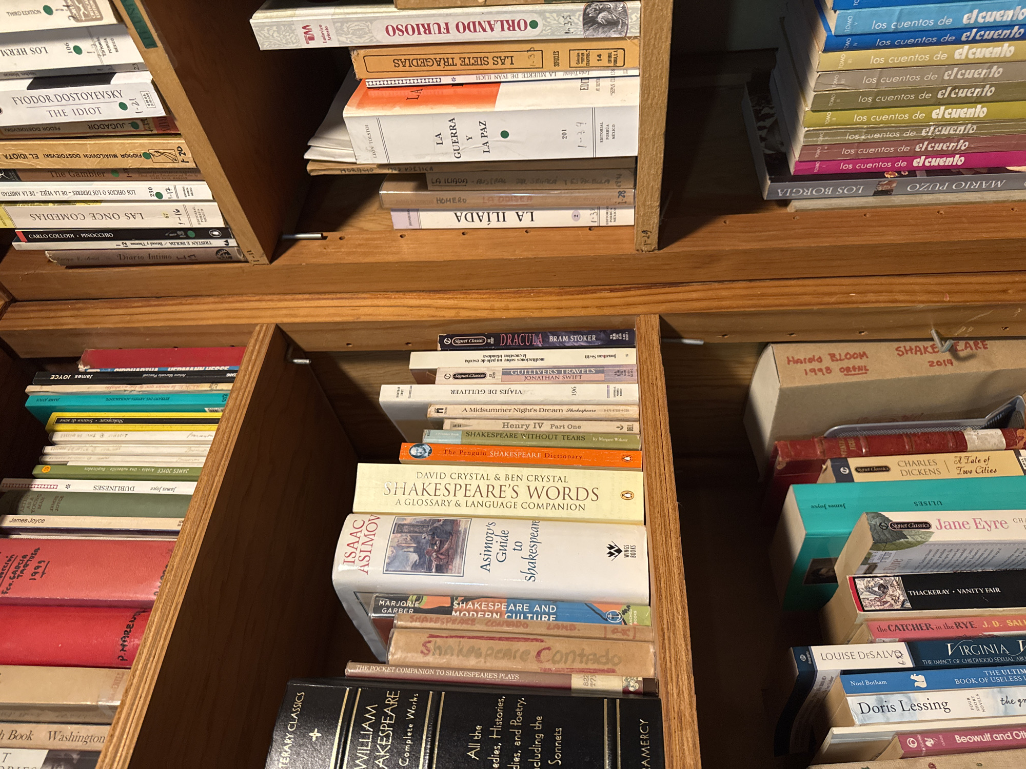 The image shows several shelves of a wooden bookcase filled with books in an indoor setting. The books vary in size, shape, and color, and many have titles in English and Spanish. Some recognizable titles include works by Shakespeare, Dostoyevsky, James Joyce, and Bram Stoker ("Dracula"). The organization of the books appears somewhat haphazard, with some stacked vertically and others horizontally. There are no people visible in the image. The environment is warm-toned due to the wood of the bookcase. There is no clear indication of the location being Mexico City; that information cannot be verified.