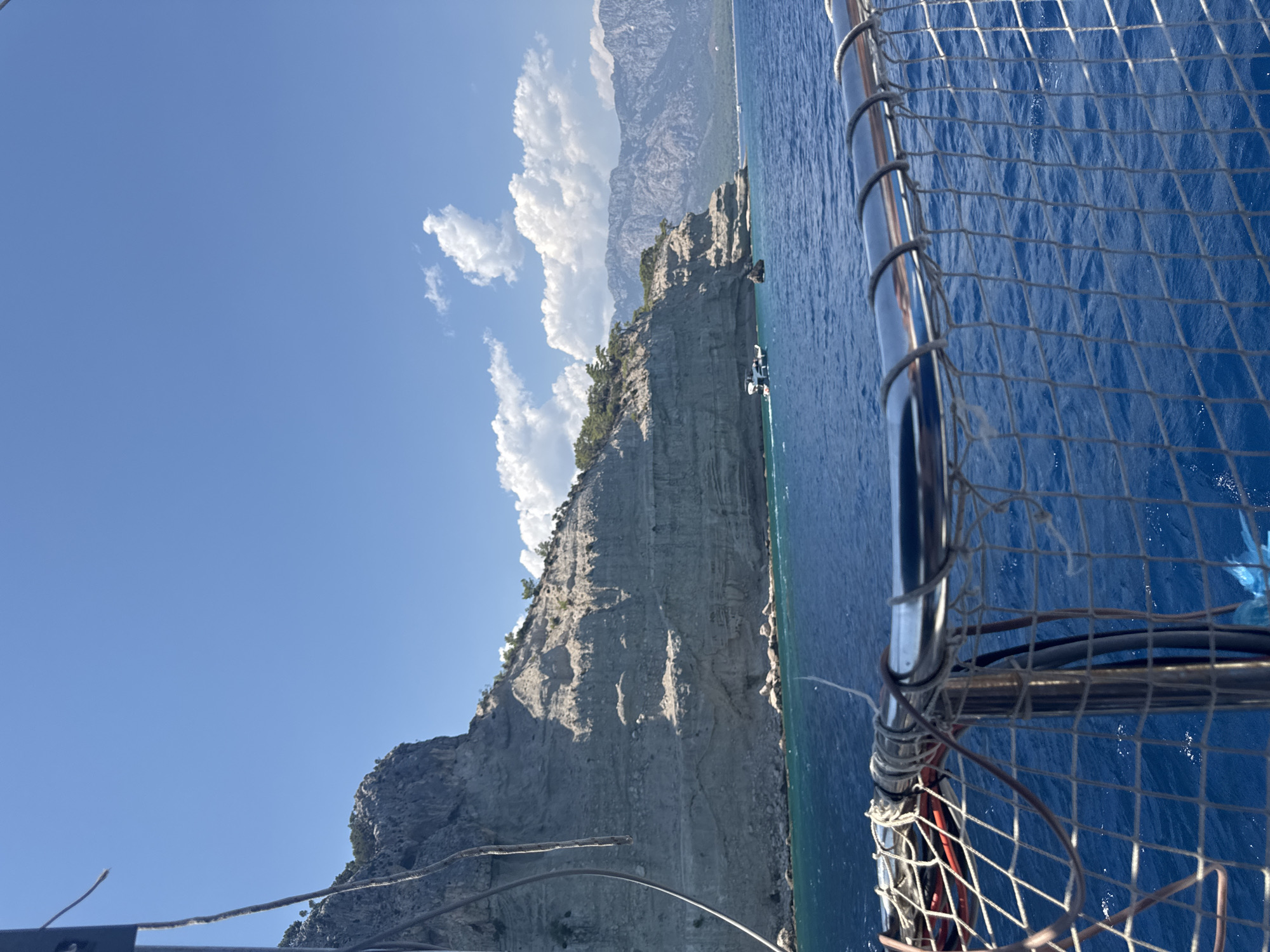 The image shows a view from a boat looking towards a rocky coastline with lush green trees on top. The sea is a deep blue, and the sky is clear with a few white clouds. In the distance, mountains can be seen. There are a couple of small boats in the water near the coastline. The foreground of the image shows a railing with netting, suggesting the viewpoint is from a boat. This scene is consistent with the coastal areas around Kemer, Turkey, known for its dramatic cliffs and clear blue waters.