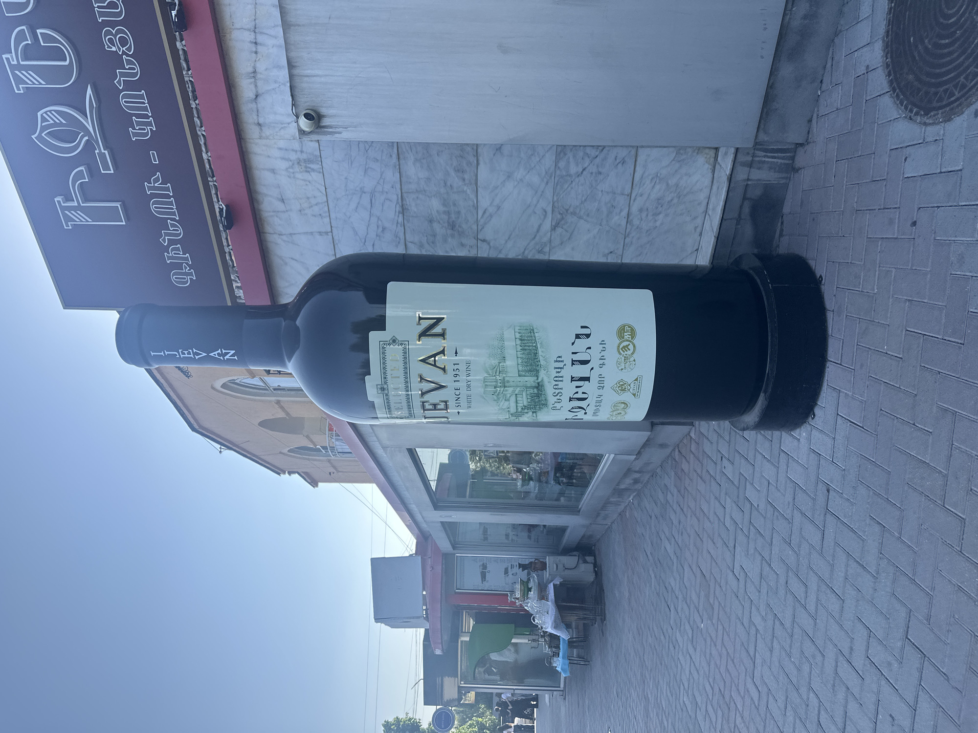The image captures a sunny outdoor scene in Gyumri, Armenia, featuring a towering, oversized replica of a dark wine bottle placed on a brick-paved sidewalk. The bottle is branded with the name "JEVAN" and has text in Armenian script. Behind the bottle, a building with a red awning and marble-patterned cladding is visible. The building has large windows, and inside, displays of what appear to be glassware and bottles can be seen. To the left of the main building, another structure with an open front displays various items on tables, including glassware and what looks like an urn. Several people are present in the background, some walking and others standing near the displayed items. The sky is clear and blue.