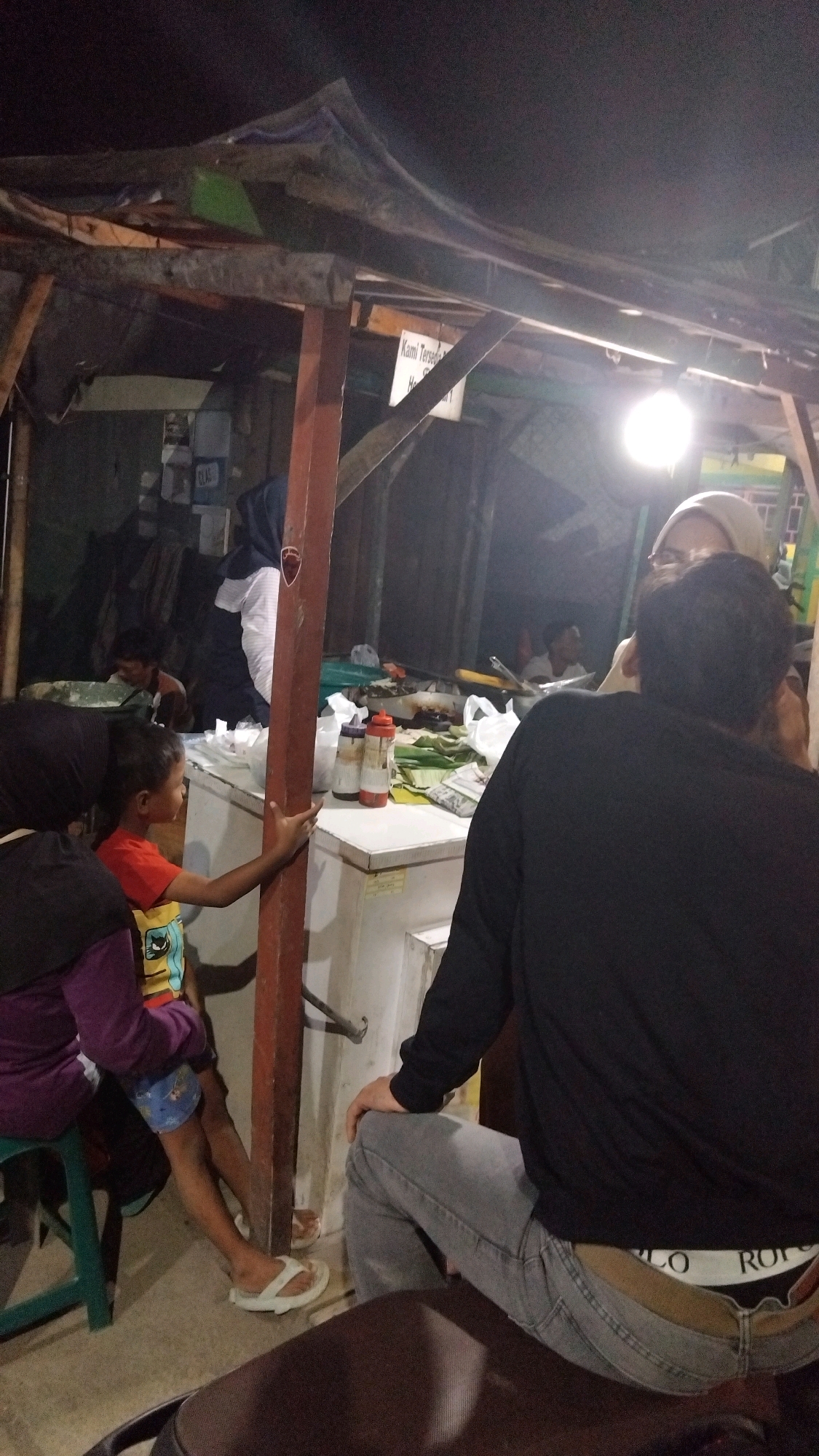 The image depicts a nighttime scene at a food stall in Tegal, Indonesia, characterized by a rustic, open-air setup with a corrugated tin roof supported by wooden beams and a red vertical pole. A bright light bulb illuminates the area, casting shadows.
In the foreground, a person wearing a black long-sleeved shirt and gray jeans is seated, facing away from the camera. The waistband of their underwear, featuring the brand "POLO," is visible. To the left of this person, a young boy wearing an orange t-shirt with cartoon characters and blue shorts is sitting on a green stool, holding onto the red pole. He is wearing white flip-flops. Next to him, another person wearing a purple long-sleeved shirt and black pants is also seated on the stool, partially obscured.
Behind the boy, a white refrigerator is positioned, with several bottles of condiments on top, including two red-capped bottles. Wrapped food items, possibly in banana leaves, are also visible on the refrigerator. Further back, a woman wearing a hijab is standing near the food stall. Other individuals are present in the background, including a person in a white shirt and a person in the distance, partially obscured by the stall's structure. The overall environment appears to be a street-side establishment, common in Tegal for evening food vending.