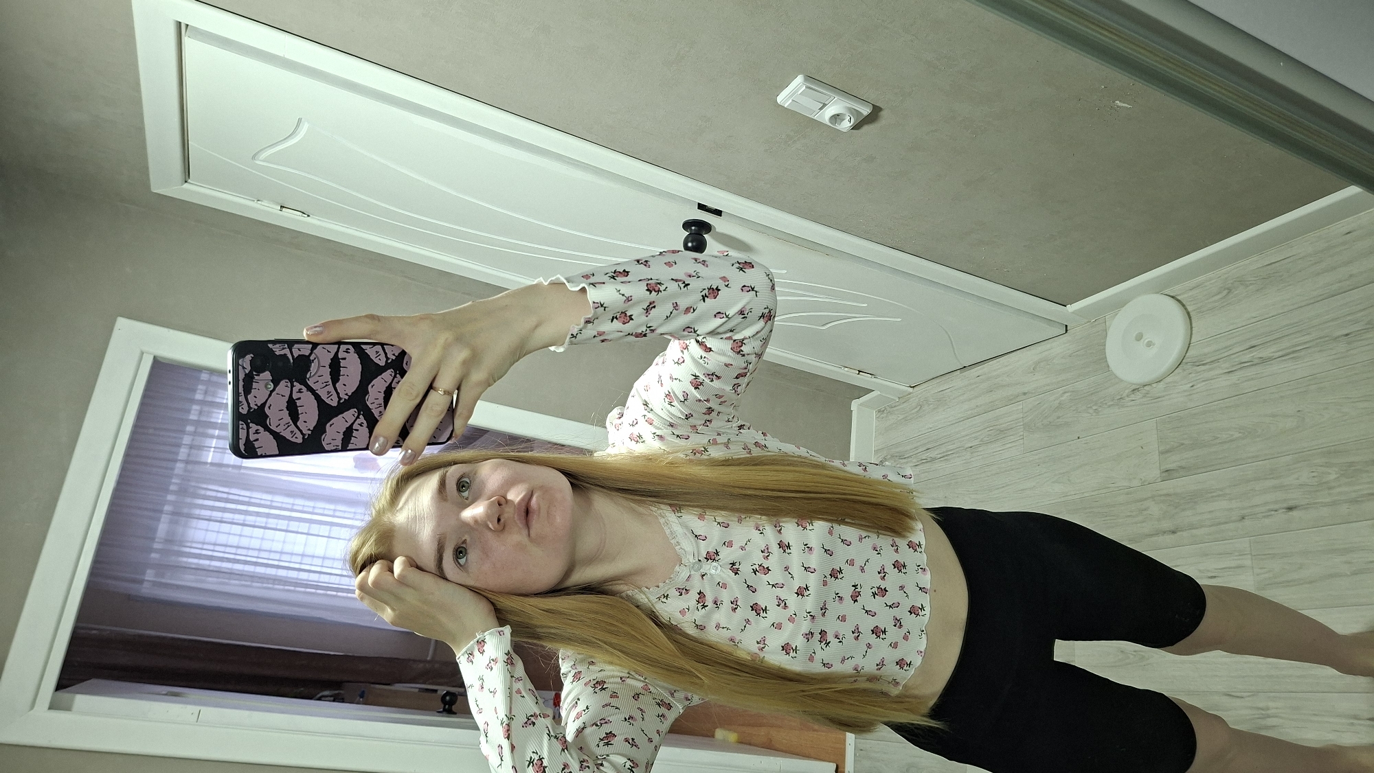A young woman with long, straight blonde hair is holding a black smartphone in her right hand, taking a selfie. The phone has a black case with a pattern of pink lip prints. She is wearing a white, long-sleeved crop top with a small floral pattern, and black bike shorts. She is lying on her left side, with her left hand supporting her head. Her face is slightly tilted towards the camera, and she has a neutral expression.
The background includes a white interior door with decorative panels, a gray ceiling with a rectangular light fixture and a white round fixture on the light wooden floor. A window frame with sheer curtains is visible to the left of the woman. The scene appears to be indoors, possibly in a residential apartment in Chaikovsky, Russia.
