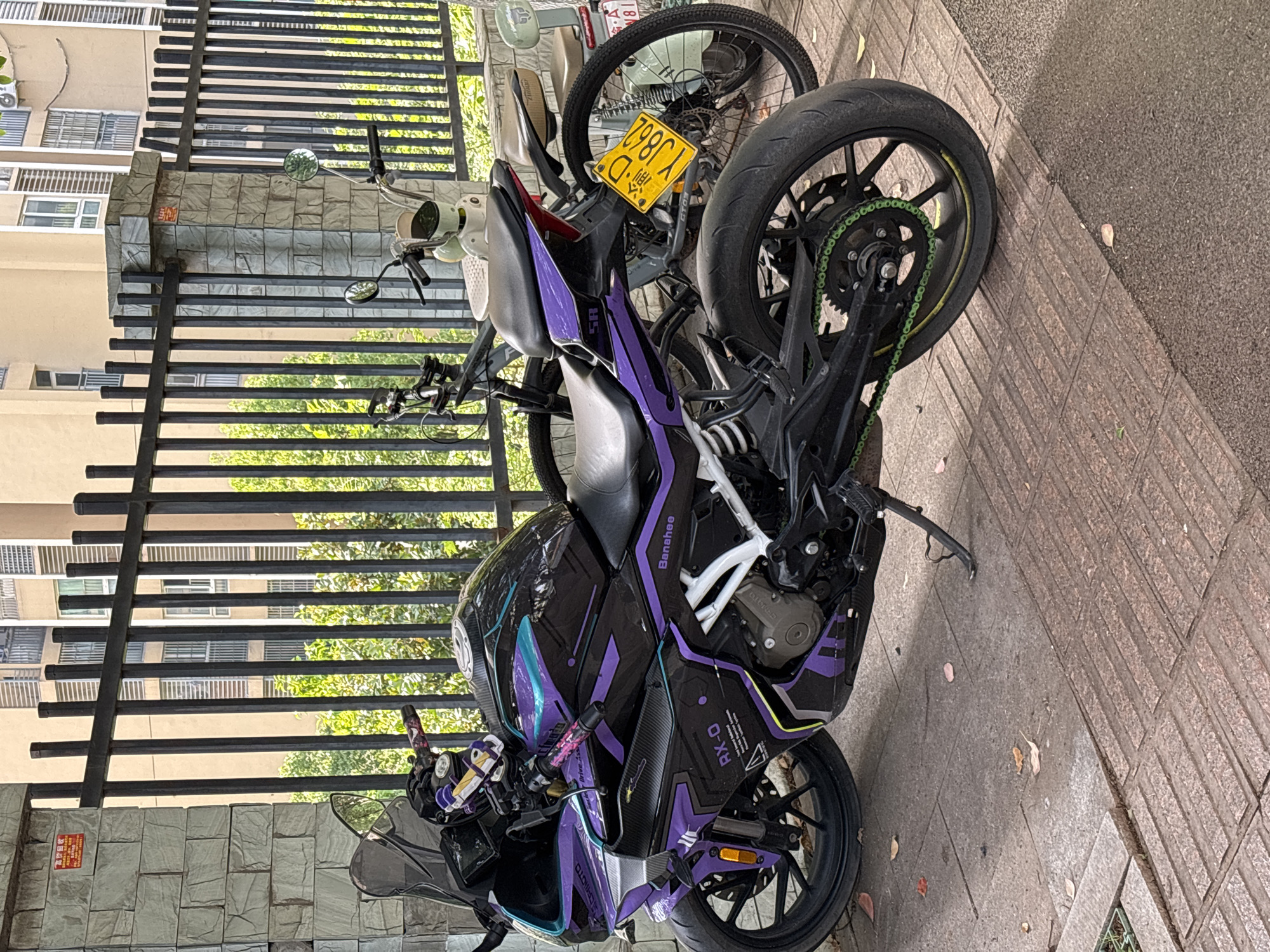 An outdoor, eye-level shot captures a custom black and purple motorcycle parked on a paved surface in Jiangjin, China. The motorcycle is the central subject, featuring a striking design with teal accents and a visible lime green drive chain. Text on the fairing includes "MOTO," "Driv," "RX-0," "Banahee," and "SA." Behind it, part of another lighter-colored bicycle or scooter is visible, with its yellow license plate displaying "渝 D YJ862," confirming its origin from Chongqing.
The motorcycle is parked next to a black metal fence, behind which residential buildings with numerous windows are visible. Lush green foliage can be seen through the fence, suggesting trees or bushes in the vicinity. The ground is a mix of textured concrete pavers and a rougher asphalt-like surface, with a few scattered dry leaves.
On a stone-clad wall behind the fence, a small red warning sign is partially visible, showing characters such as "高温" (high temperature) and hinting at a safety message like "注意安全" (pay attention to safety). The scene is brightly lit, indicating daytime. No people are present, and the vehicles appear to be stationary.