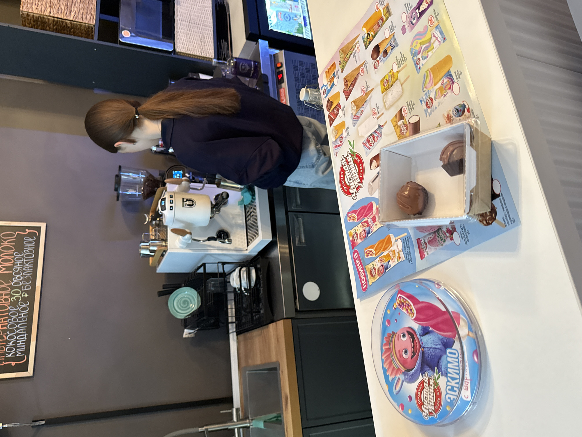 A person with long brown hair pulled into a ponytail, wearing a dark blue top and light blue jeans, stands behind a coffee machine in a dimly lit cafe. The person is seen from the side, looking down and to the left. The cafe appears to be modern, with dark gray walls and wooden accents. On the counter are various items, including a coffee grinder, a white coffee machine with a portafilter, and a black metal rack holding plates and a green saucer.
To the right of the coffee machine, there is a display of ice cream products. These include several ice cream bars and cones with colorful packaging. A small cardboard box displays two chocolate-covered ice cream treats, one of which has been partially eaten. A circular container of "Эскимо" (Eskimo) ice cream, featuring a cartoon character, is prominently placed in the foreground. Some visible text on the packaging is in Russian, mentioning "Чистая Линия" (Chistaya Liniya - Clean Line) as a brand, and "Эскимо" (Eskimo). Other text indicates different ice cream flavors, such as "кокосовое" (coconut), "миндальное" (almond), and "безлактозное" (lactose-free). There is also a chalkboard sign on the left wall with handwritten text in Russian, listing milk, coconut, almond, and lactose-free options. The overall scene suggests a typical day in a cafe or ice cream shop.