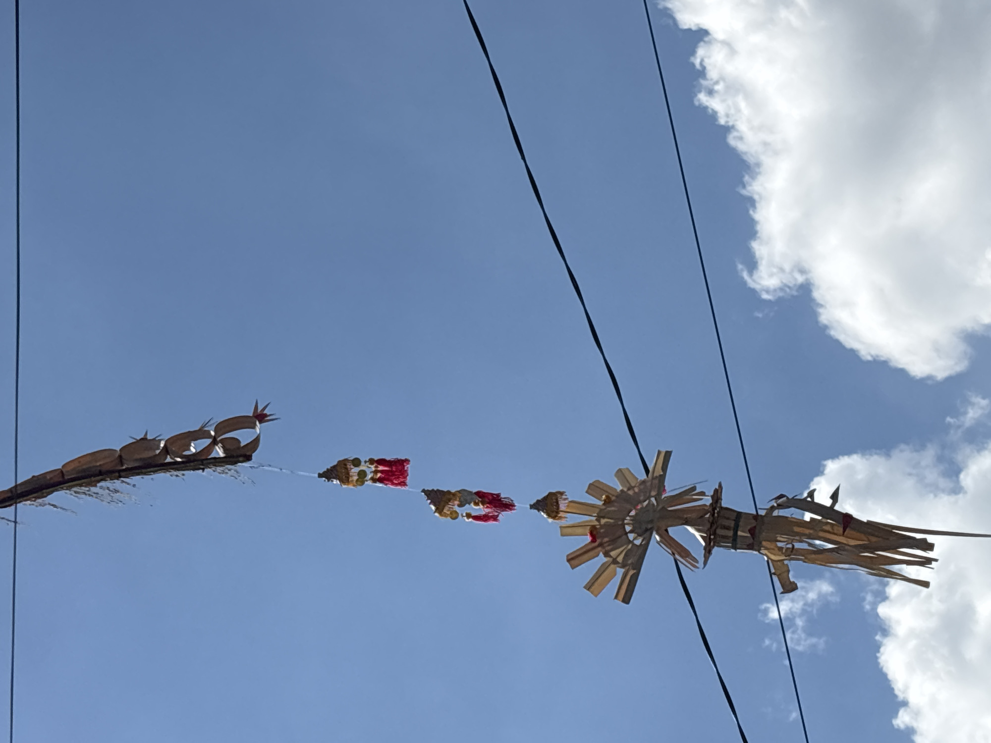A hanging, traditional Balinese "penjor" made of woven palm leaves and adorned with golden, bell-shaped ornaments and red tassels is suspended from a wire against a clear blue sky with scattered white clouds. The penjor is in the process of being decorated, as indicated by its unfinished appearance and dangling decorative elements. The scene is outdoors, suggesting it is part of a celebration or religious ritual in Mengwi, Indonesia. The wires appear to be electrical or telephone lines.