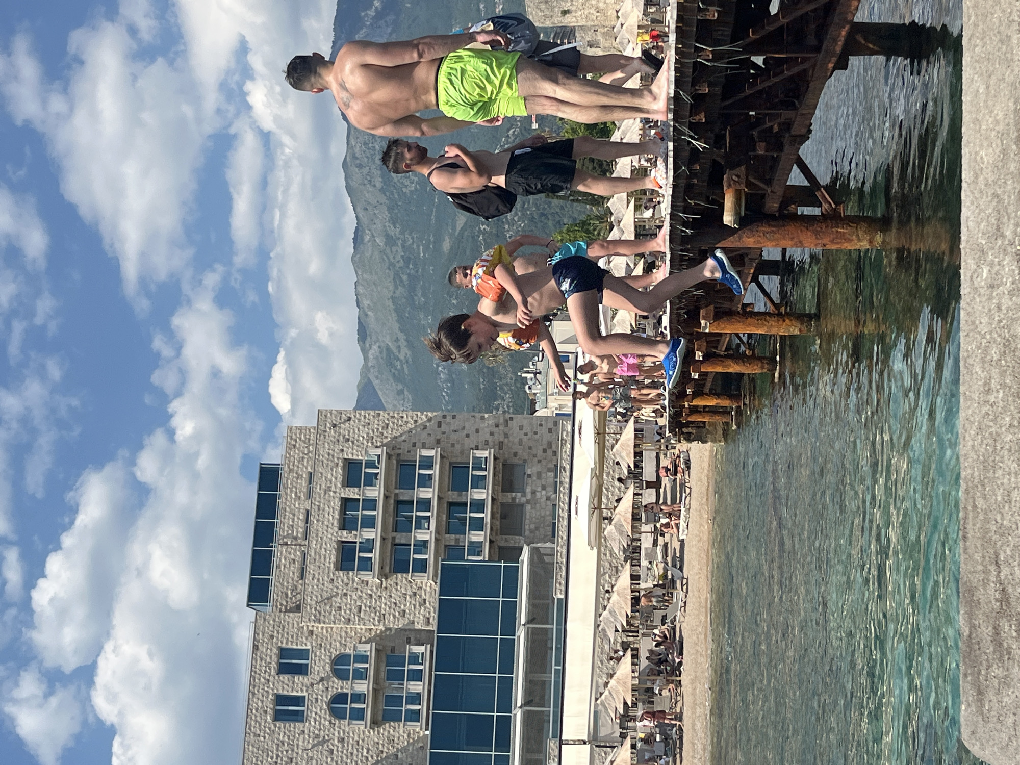 In this sideways, natural light photograph, a group of people are enjoying a sunny day by the water. The scene appears to be a lively beach or waterfront area, with a modern, multi-story building constructed of light-colored stone and dark blue windows as a backdrop on the left. A rusted metal pier extends into the clear turquoise water on the right, where several individuals are visible, some standing near the edge and others interacting with each other.
Several people are in swimming attire. One man in lime green shorts is standing, his back to the camera, looking out towards the water. Another man in black shorts is standing next to him, also facing away. Further down, a younger person in blue swim shorts is in mid-air, appearing to be jumping into the water, while another child in dark swim trunks is close by, possibly about to follow. There are other individuals scattered along the pier and on the beach, some relaxing on lounge chairs under umbrellas.
The weather seems to be pleasant, with a bright blue sky dotted with fluffy white clouds. The sun is shining, casting reflections on the water and highlighting the textures of the stone building and the rust on the pier. The overall mood is one of leisure and recreation, suggesting a holiday or a relaxed outing. No visible text is present in the image.