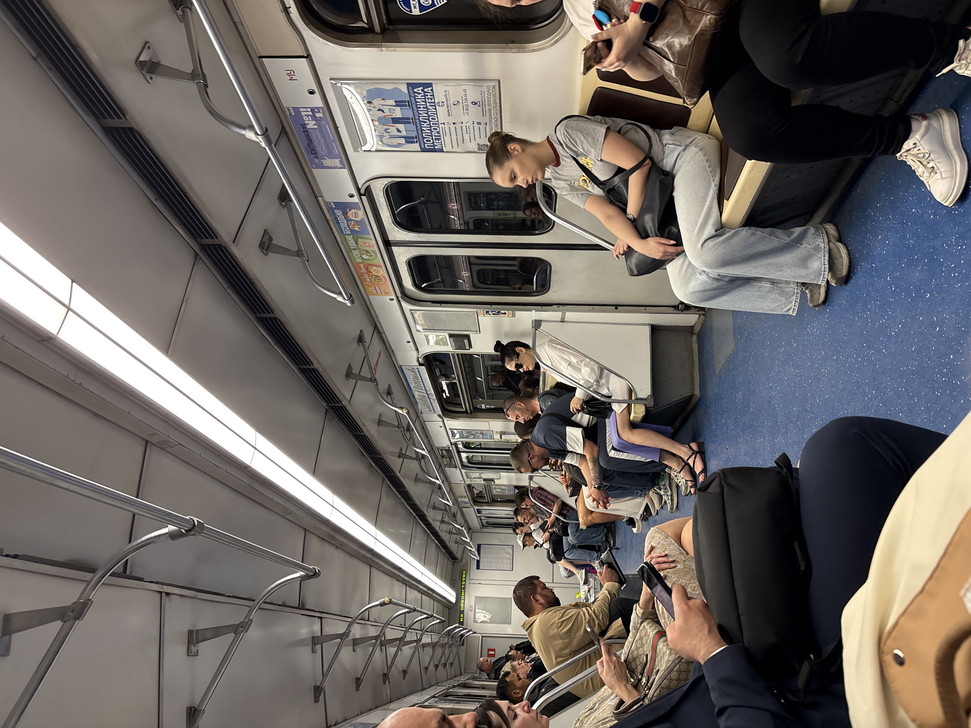 The image shows the interior of a subway car in Saint Petersburg, Russia. Several passengers are seated or standing, most are using their mobile phones. The car has blue flooring and light-colored walls with advertisements. Overhead are light fixtures and handrails. Some advertisements include healthcare information for the metro, and food product information.