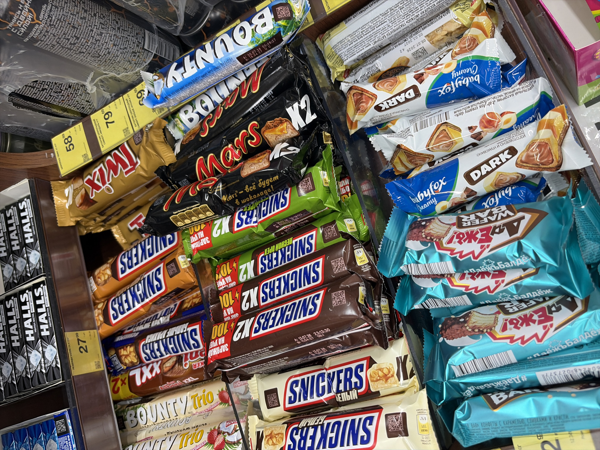 The image shows a shelf in a store, likely a supermarket or convenience store, filled with various chocolate bars and candy. The products are arranged in rows and packs, with some individual bars visible. Among the brands identifiable are Snickers, Mars, Twix, Bounty, and Halls. The packaging is colorful and features brand logos and product images. Some of the products are in their original wrappers, while others are displayed more loosely. The lighting suggests an indoor setting, and the overall impression is of a commercial display of confectionery items. The presence of Cyrillic text on some of the packaging, such as on the Halls boxes and some of the chocolate bar wrappers, indicates a Russian-speaking market. Without further contextual information or visible landmarks, it is impossible to definitively place this image in Moscow, Russia, or any other specific location. The scene depicts a retail environment focused on selling sweets.