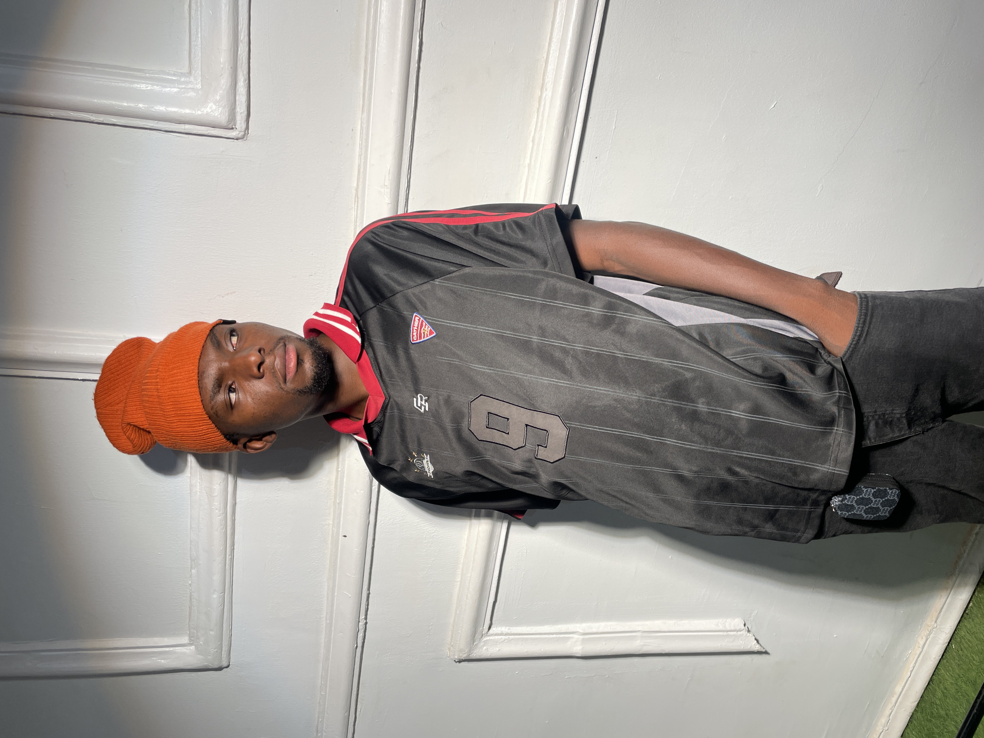 A person stands indoors, leaning against a white paneled wall. The person wears an orange beanie, a black jersey with the number 6 on it, and dark pants. The person's hands are behind them. A small, patterned pouch hangs from the person's pants. Green turf is visible at the bottom right of the frame. There is no indication of location, city, or environment.