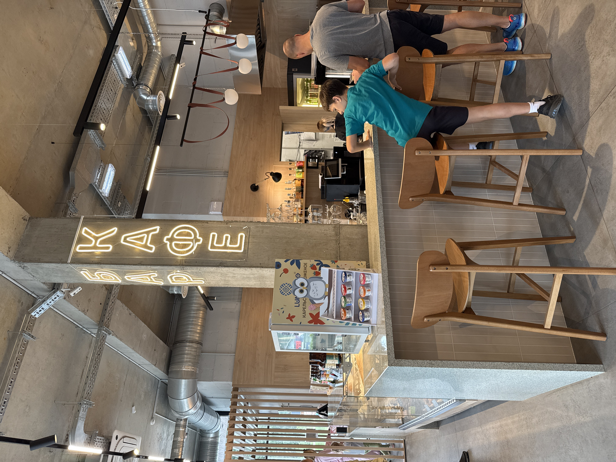 The image depicts the interior of a modern cafe with an industrial aesthetic. A concrete ceiling with exposed ductwork and lighting fixtures hangs above. A large concrete pillar in the foreground is adorned with bright neon signage that reads "БАР КАФЕ" in Cyrillic letters. The cafe counter is made of speckled granite with a backsplash of light grey tiles. On the counter, a small refrigerated display case contains several colorful tubs of ice cream, with a prominent display of a cartoon owl.
To the right of the counter, two individuals are standing. A man wearing a grey t-shirt and black shorts is on the right. A young boy in a teal t-shirt and black shorts is leaning on the counter, facing away from the camera and towards the man. Both are positioned near wooden bar stools. In the background, shelves behind the counter hold bottles and glassware. The floor is a polished grey concrete. The overall scene suggests a casual establishment, likely a cafe or ice cream parlor, in Yakkima, Russia.