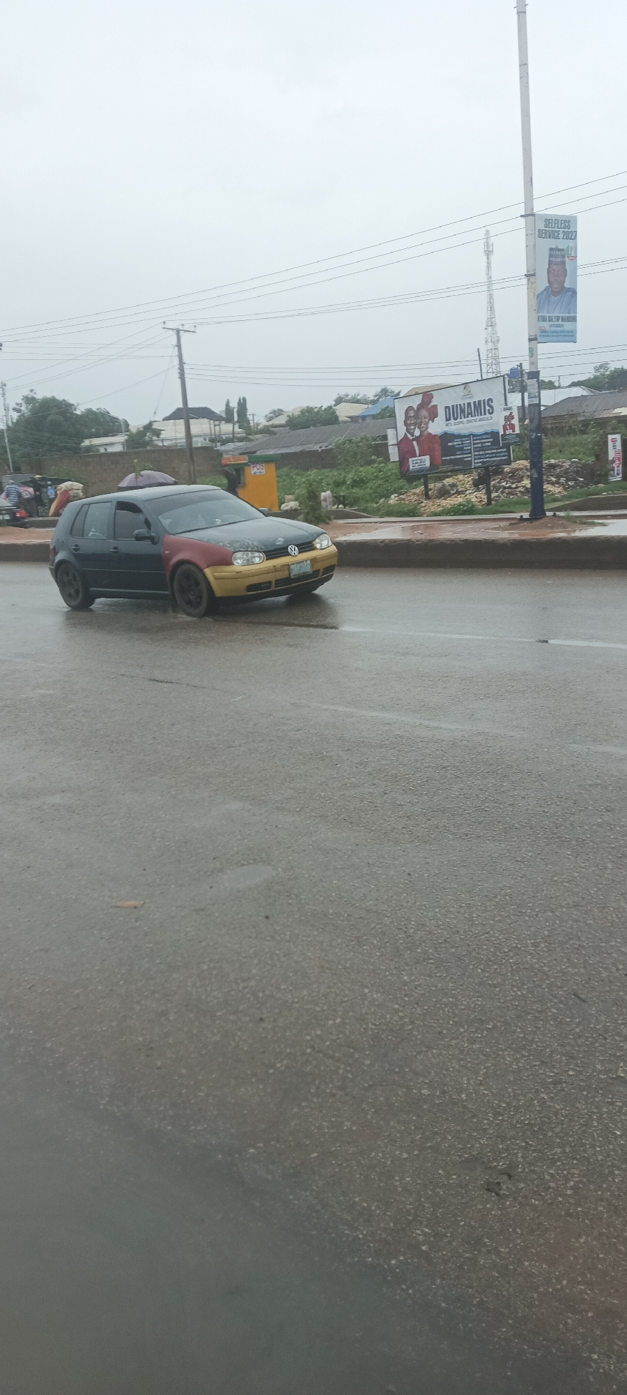 This image captures a street scene in Bukuru, Nigeria, likely during or immediately after rainfall, given the wet asphalt road and overcast sky.
The primary subject is a multi-colored Volkswagen Golf car, appearing to be in motion. Its body is black, the front passenger-side fender is red, and the front bumper and a portion of the hood are yellow. The car has dark rims. A person is partially visible inside the car through the rear window, and an umbrella is seen near the vehicle, suggesting another person might be sheltering from the rain.
The setting is an urban or semi-urban area. The wet, grey asphalt road occupies the foreground and midground. To the right of the road, there's an unpaved shoulder with some green vegetation and what appears to be a concrete barrier or curb. In the background, various buildings, fences, and utility poles are visible.
Notable objects include two prominent billboards. On the right side, a tall pole displays a banner with the text "SELFLESS SERVICE 2027" and a portrait of a man, with the name "ATIRA BALTIP MANDUN" partially visible. Further back, another billboard advertises "DUNAMIS INT'L GOSPEL CENTRE, ABUJA," featuring images of a man and a woman smiling. Utility poles with electrical wires crisscross the upper portion of the frame, and a white telecommunications tower is visible in the distance. The overall impression is a bustling street on a gloomy, rainy day.