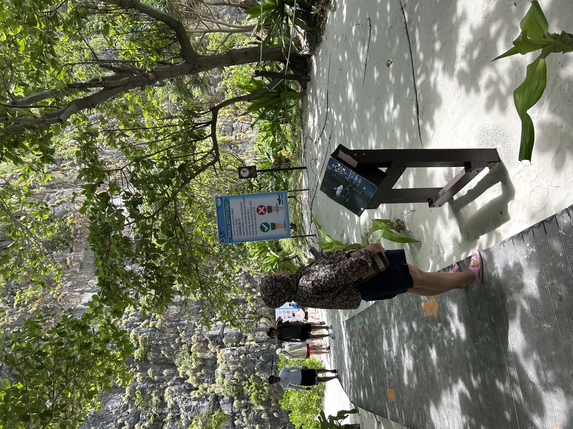 A group of people are walking on a wooden pathway in a tropical environment. The pathway is bordered by white sand and lush green vegetation, with a large, rocky cliff face covered in plants in the background. A sign with text and icons is visible, indicating "ANKLE DEPTH ONLY" with a checkmark symbol and a prohibition symbol. Another display board with images, possibly of marine life, is also present. The lighting suggests a sunny day, with shadows cast by trees and foliage. The overall scene appears to be a tourist attraction or a scenic walk, likely in Ao Nang, Thailand, given the tropical setting and prevalence of such attractions in the area.