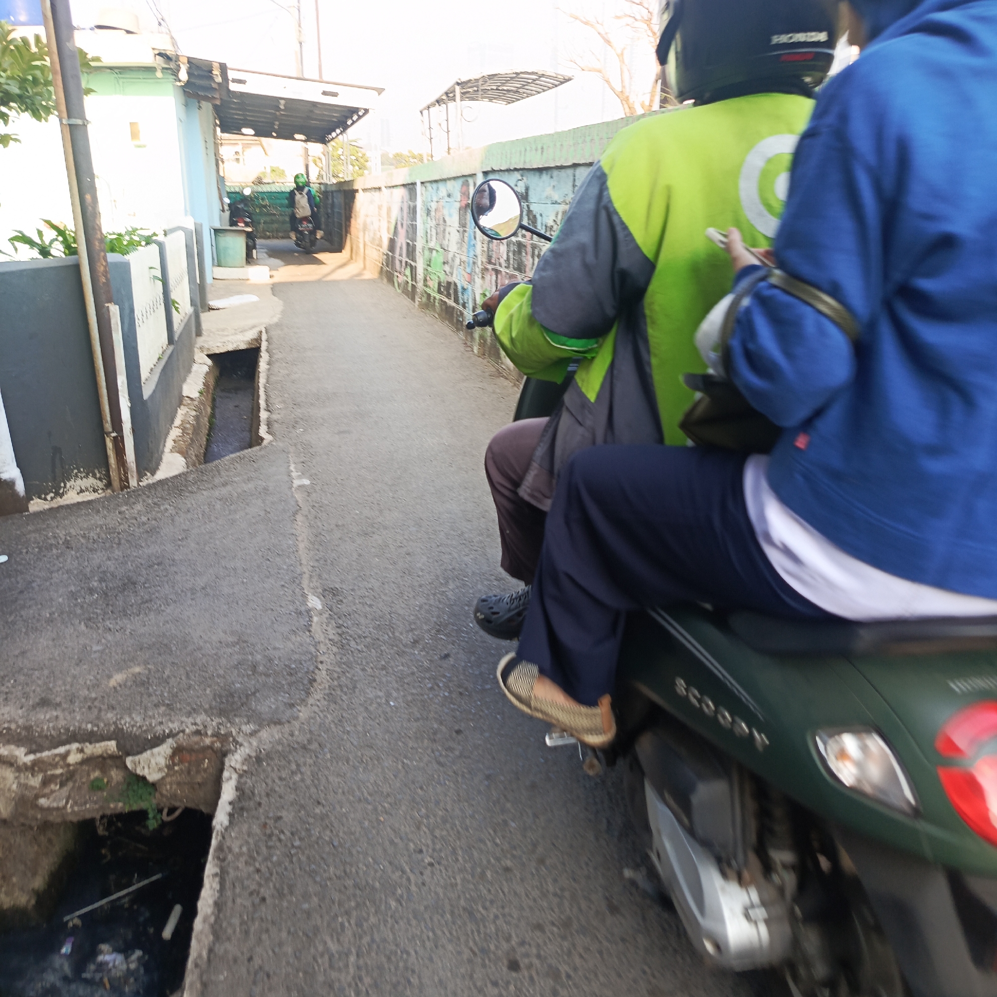 This is a low-angle, wide shot of two people on a dark green scooter, with the rider wearing a green vest and a black helmet, and the passenger wearing a blue hoodie. The scooter is moving along a narrow, paved road in what appears to be an urban residential area in South Jakarta, Indonesia. To the left of the road is a partially damaged concrete drainage ditch with murky water and some debris. Adjacent to the ditch is a wall with a dark grey lower section and a white upper section with some decorative elements. Further left, a light blue building with a white roof and a grey awning is visible. To the right of the road is a low wall with some graffiti. The road surface is asphalt with some lighter colored markings. In the background, a person on a scooter with a green helmet can be seen further down the road, and there are buildings and trees. The lighting suggests it is daytime, possibly late afternoon, with the sun casting some shadows.