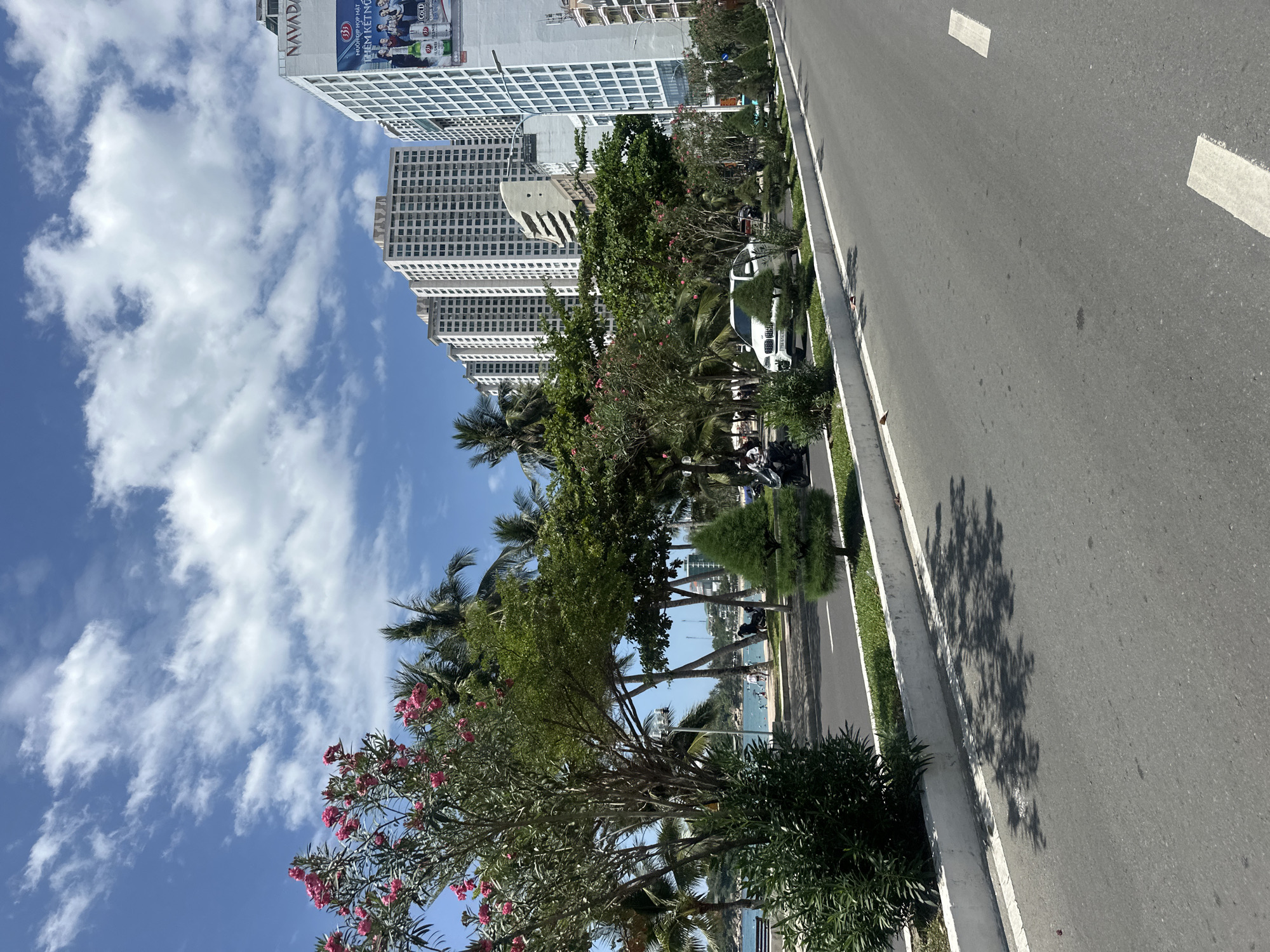 The image captures a vibrant, sunny day in an urban coastal setting, likely in Bac Nha Trang Ward, Vietnam.
On the left, several modern, high-rise buildings with repetitive grid-pattern windows dominate the skyline against a bright blue sky filled with scattered white clouds. One prominent building features a large billboard advertisement. The billboard clearly displays the brand "NAVADA" in red letters. Below it, Vietnamese text "MƯƠI HƠN MẤY" and "DÒNG KẾT NỐI" is visible, alongside images of beer cans (including faint "Coors Light" and "MILLER LITE" logos) and people toasting, suggesting an advertisement for a beverage or a celebration.
A wide, multi-lane asphalt road occupies the right side of the frame, marked with white dashed and solid lines. A distinct, intricate shadow of a tree is cast across the road, indicating strong sunlight from a high angle.
Separating the road from the buildings is a lush, tree-lined median or sidewalk. This green strip features various trees, including tall palm trees and other deciduous trees with vibrant pink blossoms, likely oleander. Beneath the trees, a white SUV is parked or driving, and several motorcycles are also visible parked along the curb. Small figures of people can be seen in the distance, near the water's edge.
Beyond the dense vegetation, glimpses of a blue water body, likely the ocean or a bay, are visible, dotted with a few boats. The overall scene depicts a bustling, warm environment under clear skies.