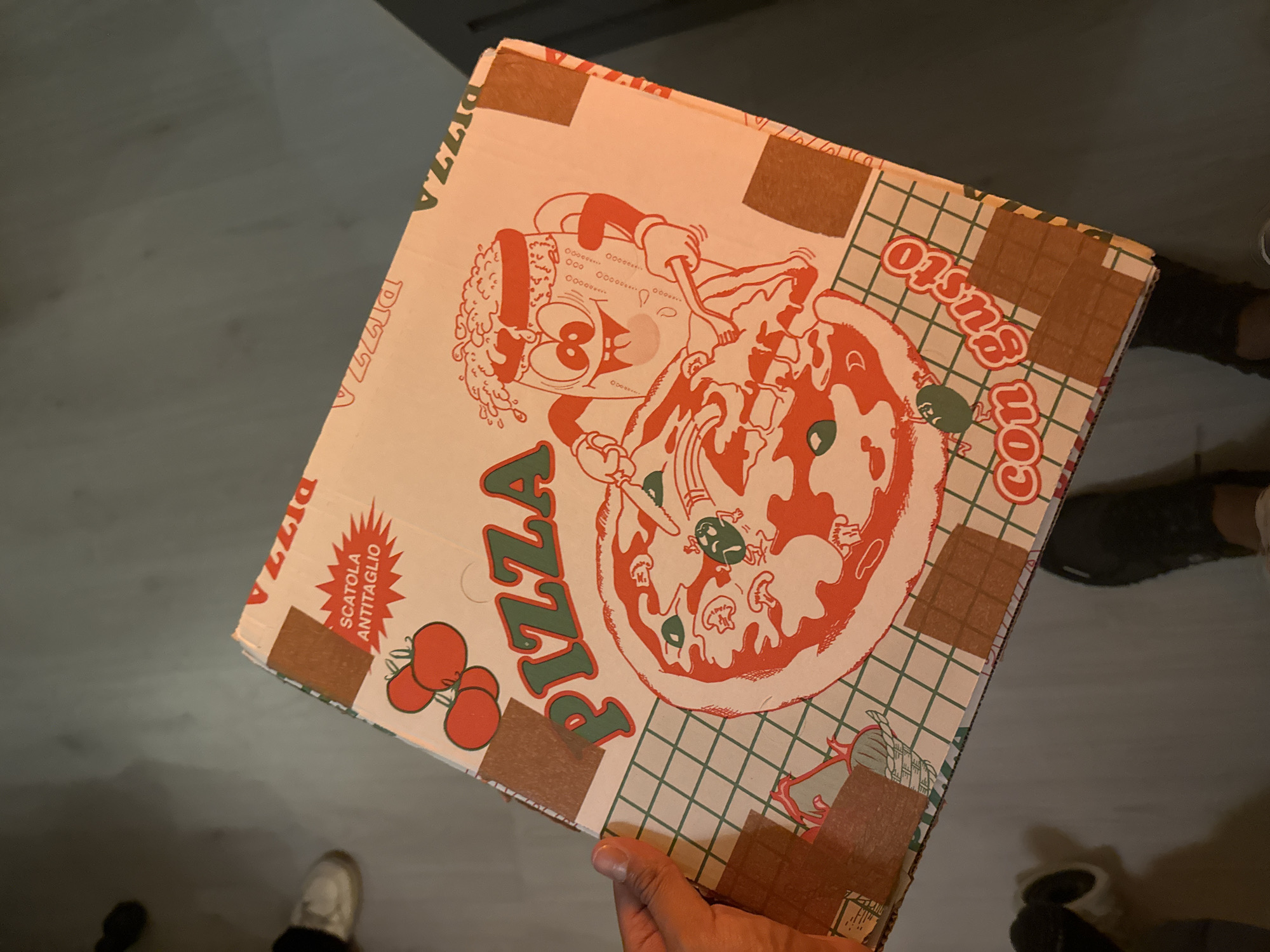 The image shows a pizza box held in a person's hand. The box is printed with a cartoon image of a chef and a pizza. The words "PIZZA" and "con Gusto" are also printed on the box. The box appears to be sealed with brown paper tape. Parts of two pairs of legs, one with white shoes and another with black shoes, and some dark spots are visible in the bottom part of the frame. The location in the city of San Pedro Cholula, Mexico cannot be determined from the image.