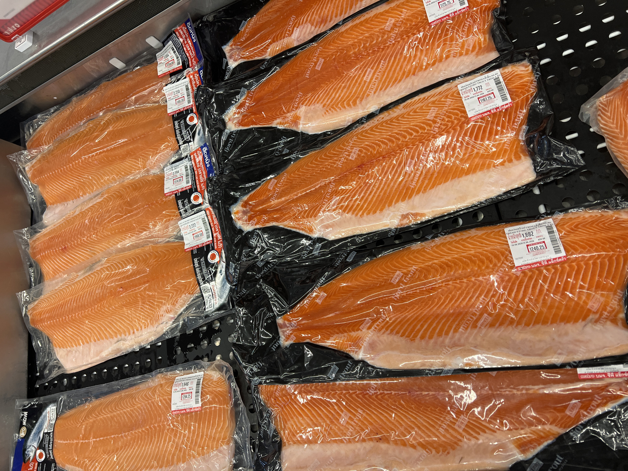 The image shows a close-up, overhead view of packaged salmon fillets displayed in a retail setting. The salmon fillets are bright orange and appear fresh, each individually sealed in clear plastic packaging. Some of the packages have white labels with text and barcodes. The fillets are arranged in rows on a black, textured surface. In the background, other rows of similar packaged fish are visible. The overall impression is of a well-stocked seafood counter. There are no people or animals visible in the image. The location is identified as Baan Bo Phut, Thailand, suggesting this is a market or supermarket in that area.
