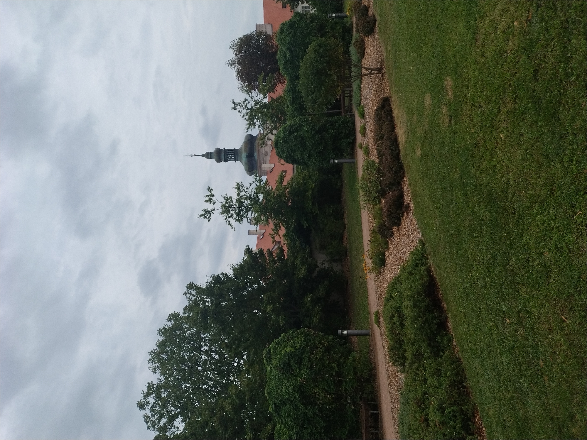 The image depicts a view of the Jesuit College complex in Kutná Hora, Czechia, under an overcast sky. In the foreground, a well-maintained garden features a gravel pathway, neatly trimmed bushes, and a lush green lawn. Several modern lampposts are strategically placed along the path. In the middle ground, a variety of trees, including some with ivy-covered branches, create a dense green backdrop. Behind the trees, the distinctive architecture of the Jesuit College is visible, characterized by its orange tiled roofs and a prominent green-domed tower topped with a cross. The overall atmosphere is serene and calm, suggesting a quiet, possibly historic, urban setting. No people are present in the image.