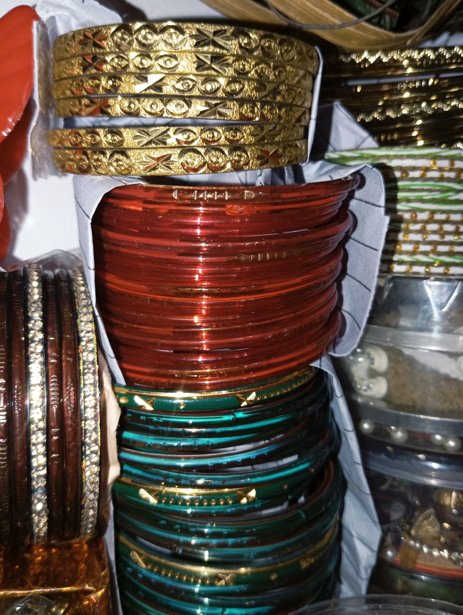 The image displays a close-up view of a collection of bangles. In the foreground, several stacks of bangles are visible. The top stack consists of golden bangles with intricate patterns. Below this, a large stack of translucent red bangles is prominently featured. To the left of the red bangles, a stack of dark brown bangles adorned with sparkling rhinestones or crystals is partially visible. Further down, stacks of green and teal bangles with gold accents are arranged. In the background, more bangles and other items are visible, some in clear plastic containers. The lighting creates reflections and highlights on the bangles. The overall scene appears to be an indoor display of jewelry, possibly in a market or shop. The location is noted as Unknown, Unknown.