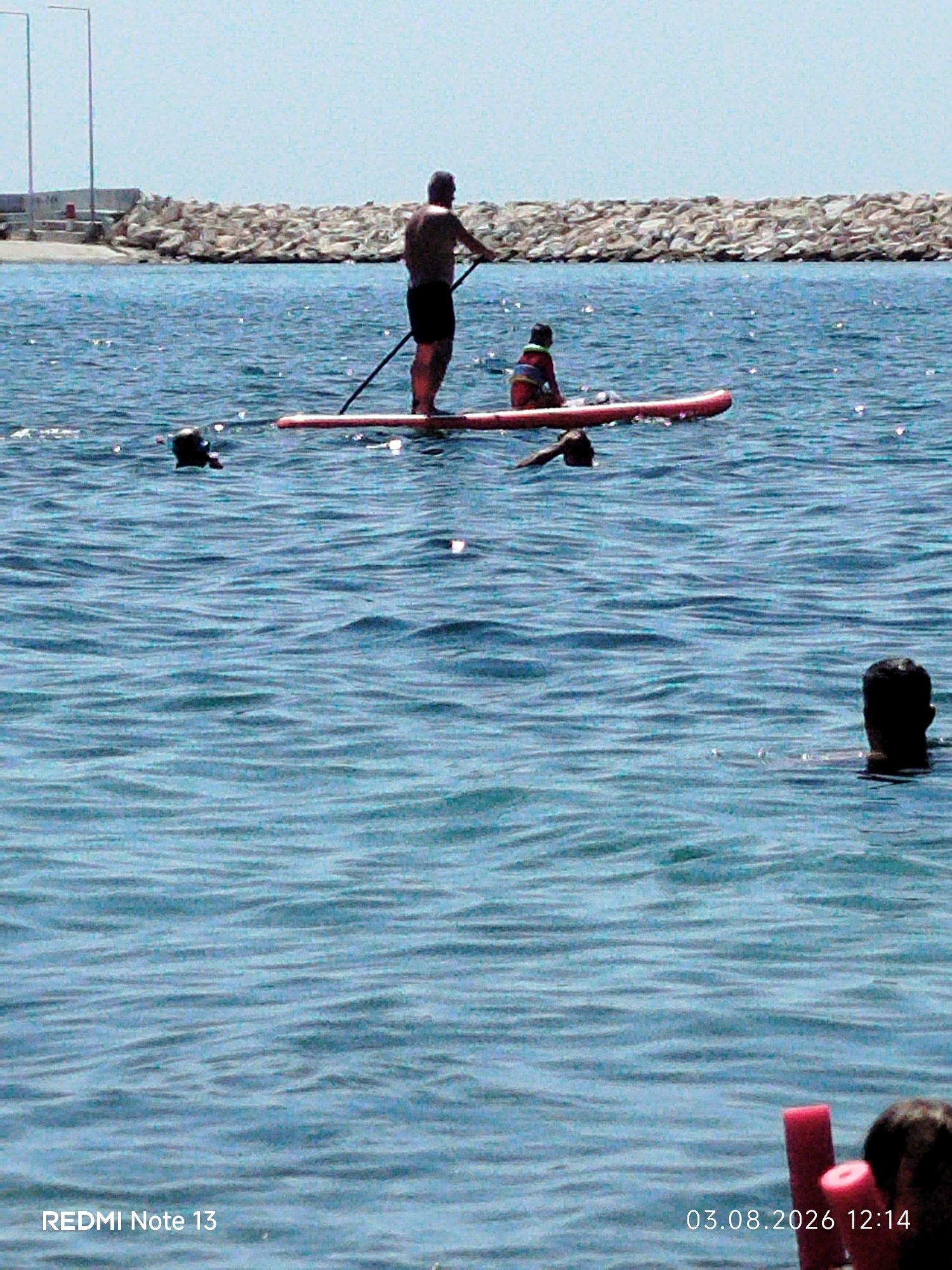 In the sunlit waters of Sithonia Municipal Unit, Greece, two individuals engage in paddleboarding. A man stands on a red paddleboard, paddling with a long oar. A child, wearing a red and white life jacket, sits on the same paddleboard. Three other people are visible in the water: one with their head above the surface, another appearing to be swimming, and a third person's head is visible near the lower right corner. In the background, a breakwater made of large rocks separates the sea from a sandy shore with two tall poles. The sky is clear and light blue.