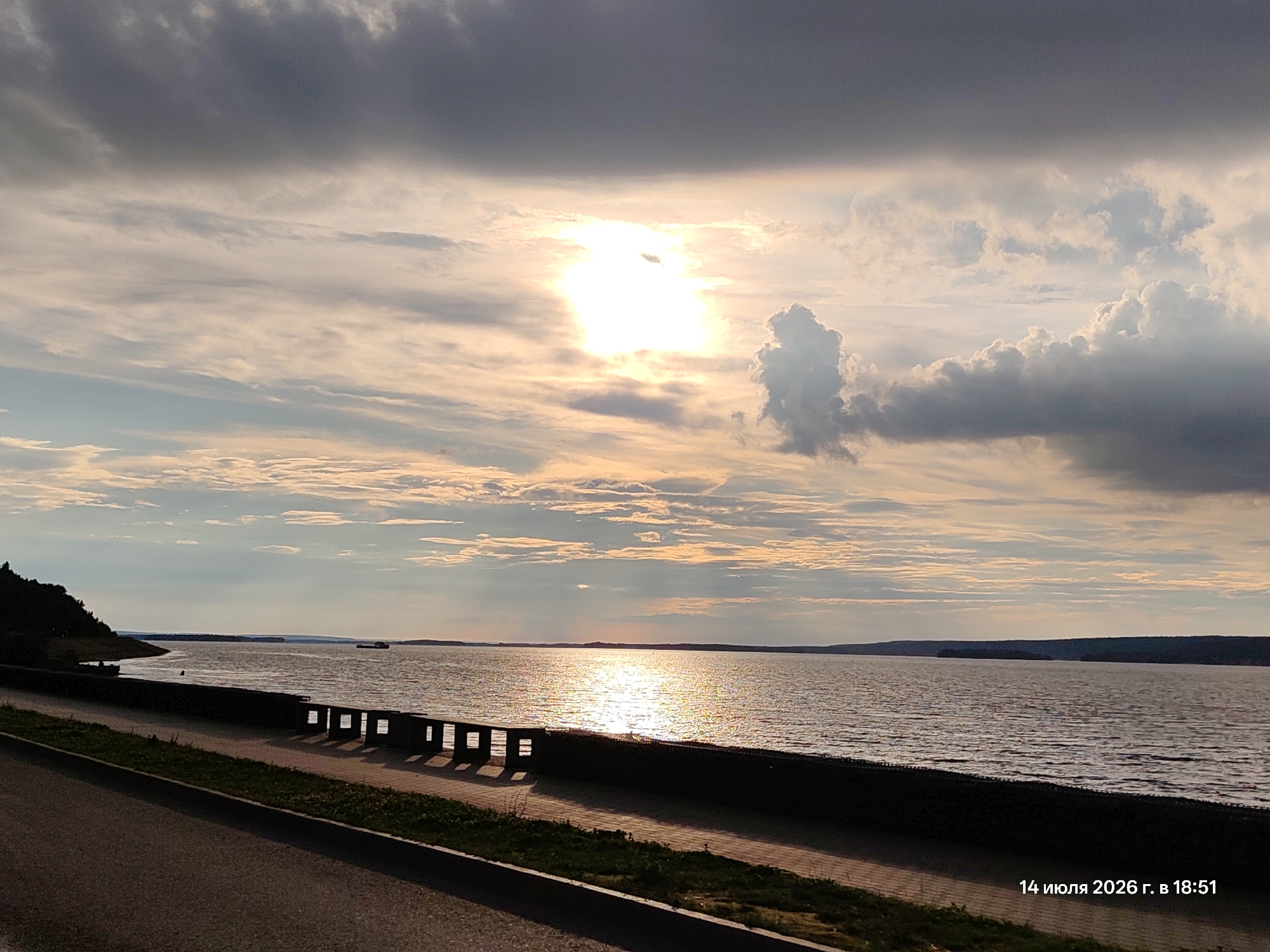 A wide shot captures a tranquil river scene at sunset. The sky is a dramatic canvas of dark, heavy clouds in the upper portion, transitioning to lighter, wispy clouds illuminated by the setting sun peeking through. The sun's bright glare reflects intensely on the rippling water, creating a shimmering path. The foreground features a paved walkway with a dark, low wall interspersed with square openings, casting long shadows. A strip of green grass borders the walkway. On the left, a dark, tree-covered hillside slopes down towards the water. In the distance, a few boats are visible on the water, and the horizon is marked by a faint outline of land. A timestamp in the lower right corner reads "14 июля 2026 г. в 18:51", indicating the date and time.