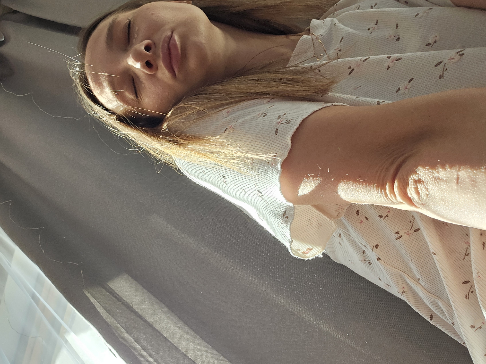 A young Caucasian woman with light brown hair is shown in a close-up shot, leaning against a gray curtain. Sunlight streams through the curtain, casting diagonal shadows across the scene and illuminating her face, arm, and clothing. She has her eyes closed, and her lips are slightly pursed. She is wearing a light-colored t-shirt with a subtle floral pattern. The background consists of the gray curtain and a portion of a window. There are fine, light-colored hairs or fibers visible on the curtain and on the woman's hair. The image appears to have been taken indoors, possibly in a residential setting. The specific location in Chaikovsky, Russia, cannot be determined from the visual information provided.