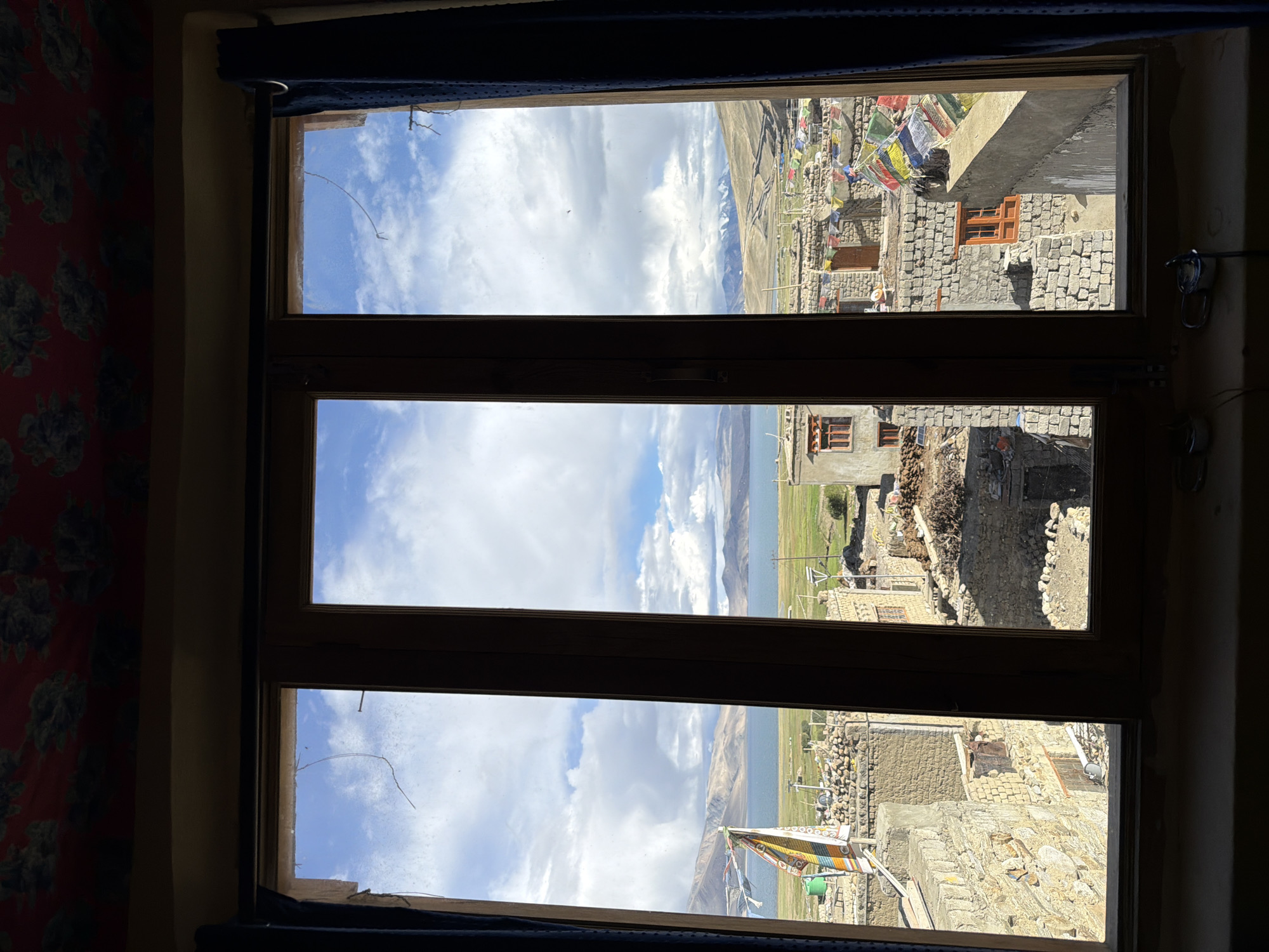 The image is a view through a three-pane wooden window of the town of Karzok, India. The window frame is dark brown, and the glass is somewhat dusty with a thin, curved crack visible in the upper left pane. Dark blue curtains are drawn back on either side of the window.
The view outside shows a village with traditional stone and mud-brick buildings, many with flat roofs. Prayer flags, colorful and rectangular, are strung between some of the buildings, indicating a Tibetan Buddhist influence. In the mid-ground, there are stacks of what appear to be animal dung, likely used for fuel. Beyond the village, a large, calm body of water, likely Tso Moriri lake, stretches out, bordered by rolling hills and distant mountains. Some of the mountains in the far distance are snow-capped. The sky is a bright blue with fluffy white clouds. The lighting suggests it is daytime, with sunlight casting shadows on the buildings and landscape. There are no people visible in the image. The foreground inside the window shows a dark windowsill with a few metallic objects.