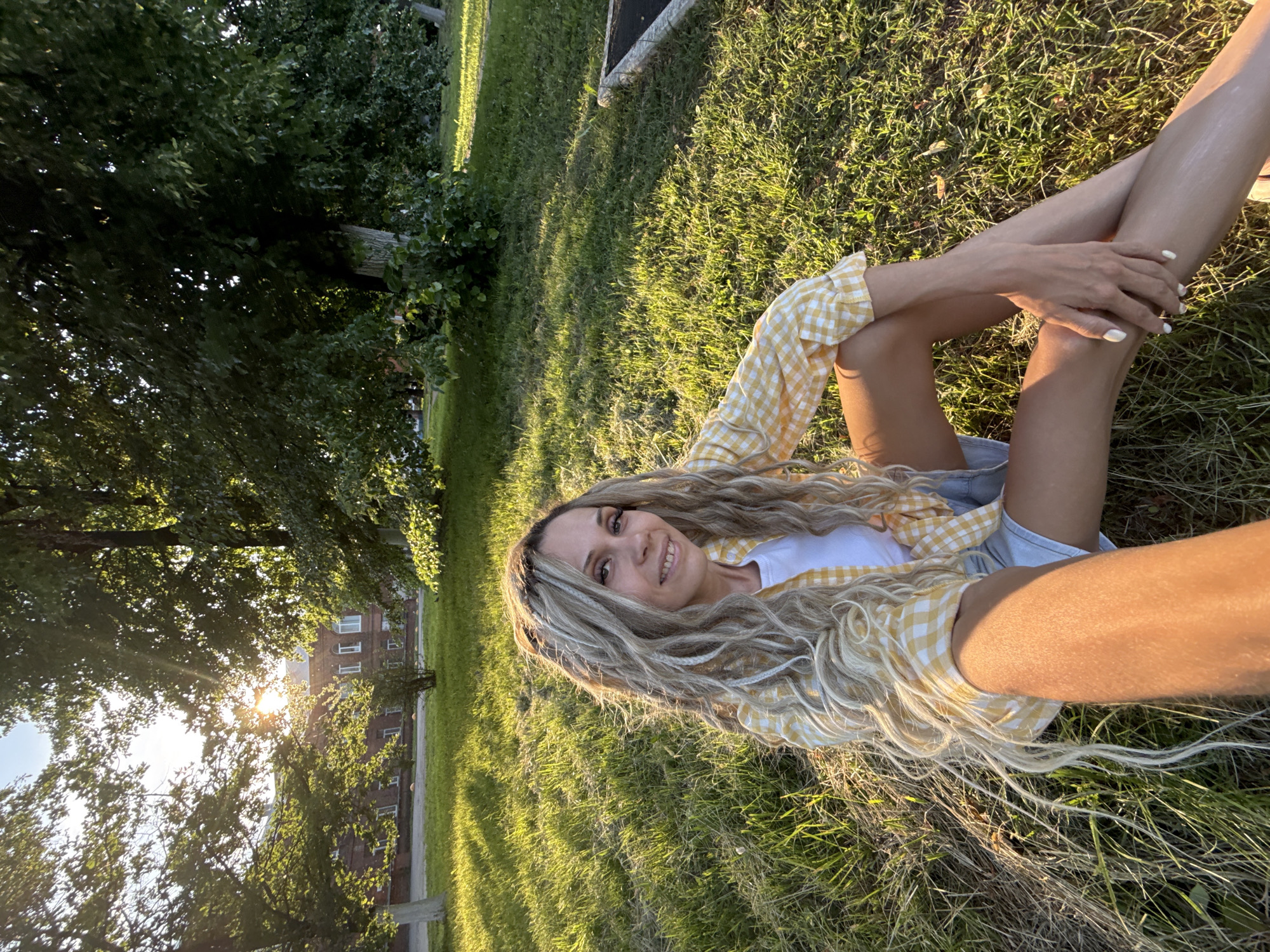 A woman with long, blonde, wavy hair is lying on a grassy lawn. She is wearing a white t-shirt and a yellow and white checkered shirt, along with denim shorts. The woman is smiling and looking towards the camera. Her legs are crossed, and her arms are resting on her legs. The sun is shining brightly, casting long shadows across the grass, indicating it is late afternoon or early evening. In the background, trees are visible, and behind the trees, a brick building with several windows can be seen. The scene suggests a peaceful outdoor setting, possibly a park or a yard. The weather appears to be warm and sunny. There is no visible text in the image.