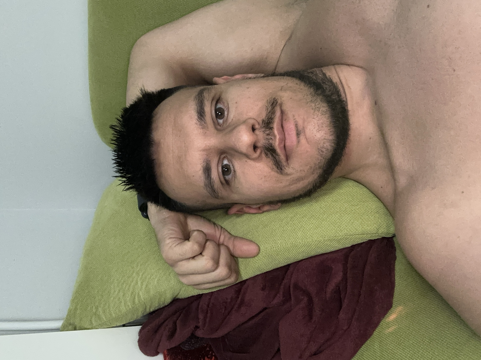 The image shows a man lying on a green and maroon pillow. He is shirtless with a beard and short black hair. His left arm is bent, and his hand is resting on the pillow. The background appears to be a white wall. No location information for Sofia, Bulgaria can be inferred from the image.
