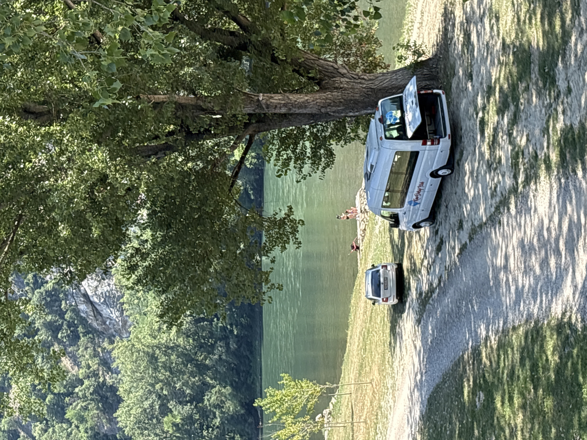 The image depicts a serene outdoor scene next to a body of water. In the foreground, a large tree with lush green foliage casts dappled shadows on the ground. Parked nearby is a white camper van with its rear door open, suggesting it might be in use or being prepared for a trip. A second, smaller white car is parked behind the van.
In the middle ground, several people are visible near the water's edge, some appearing to be relaxing or wading in the shallows. The water itself is a calm, green hue, reflecting the surrounding environment. The background reveals a lush, tree-covered hillside that rises steeply from the water, with hints of rocky outcrops.
The lighting suggests it is a sunny day, possibly late morning or afternoon, given the long shadows. The overall atmosphere is tranquil and recreational, hinting at a day spent enjoying nature by the water.
Visible text on the side of the camper van appears to be a logo with some lettering, but it is not entirely legible. The license plate on the smaller car is also too blurry to read. The location is Zvornik, Bosnia and Herzegovina, which aligns with the mountainous terrain and river setting.