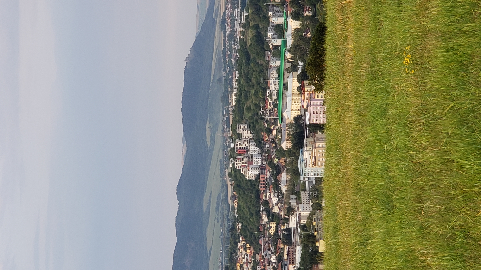 An aerial, eye-level, medium shot captures a panoramic view of a city, Prešov, Slovakia, with a lush green field in the foreground and rolling hills and mountains in the background. The city, densely packed with various buildings, including residential complexes with multiple stories and some low-rise structures, is nestled amongst rolling green hills covered with trees. The sky above is overcast, with a pale gray hue. In the distance, a prominent mountain range with a rocky outcrop on one of its peaks is visible, with lighter green fields and more scattered development on its lower slopes. The foreground is dominated by a field of tall, dry grass with a few scattered yellow wildflowers. The image appears to be taken from an elevated position, looking down on the city and its surroundings.