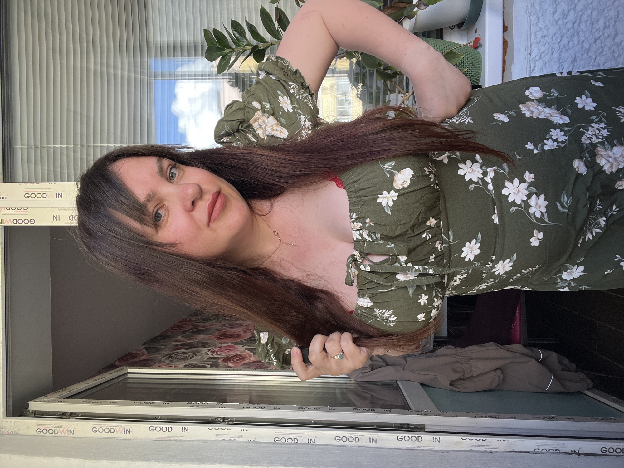 A woman with long, dark brown hair and bangs stands on a balcony, looking at the camera. She is wearing a green, floral-print dress with puffy short sleeves. Her left hand is holding a strand of her hair, and her right arm is bent at the elbow with her hand on her hip. The background shows a window with blinds, a potted plant, and floral wallpaper. The balcony has white window frames with "GOODWIN" printed on them. A brown jacket is hanging on the wall to the left of the frame. The scene appears to be in Noviy, Russia.
