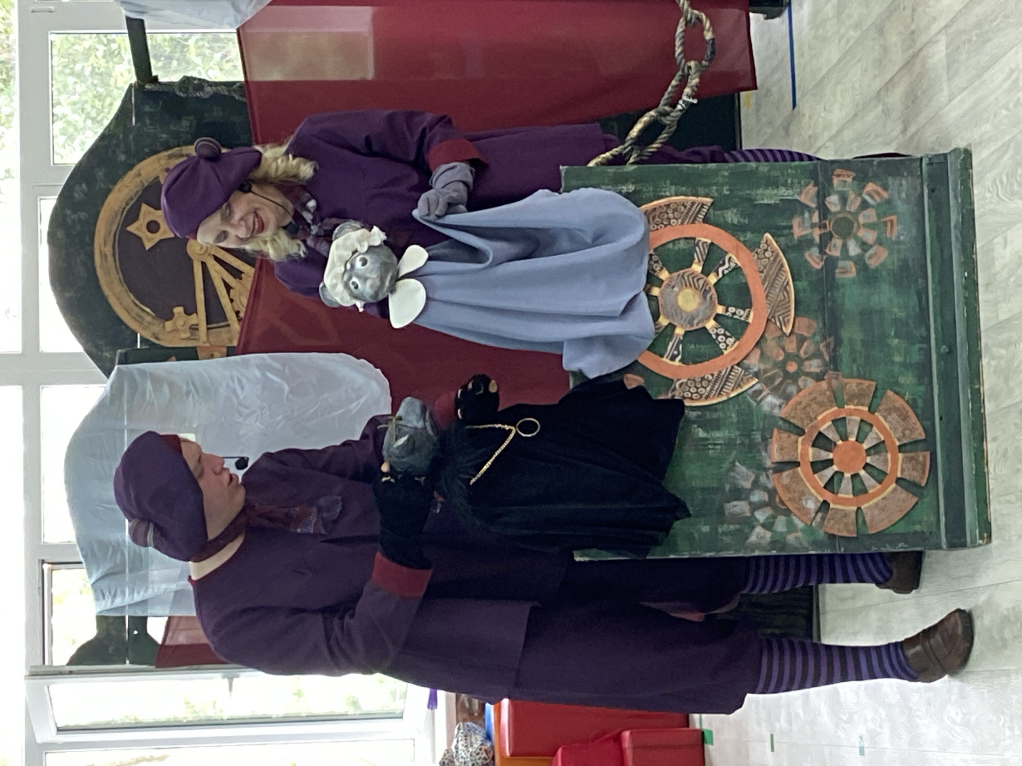 In an indoor setting, two individuals in matching purple outfits and berets are operating puppets. The individual on the left, wearing a maroon scarf and striped socks, holds a dark-colored puppet with a gold chain around its neck. The individual on the right, with blonde hair, holds a lighter-colored puppet draped in a blue fabric. Both puppets have grey, animal-like heads with white accents. They are positioned in front of a green wooden prop decorated with painted gears and metallic accents. The background features red fabric drapes, a large circular wooden structure with star detailing, and large windows with natural light entering. Stacks of red and patterned boxes are visible to the left, and a rope is seen to the right. The floor appears to be light-colored wood planks.