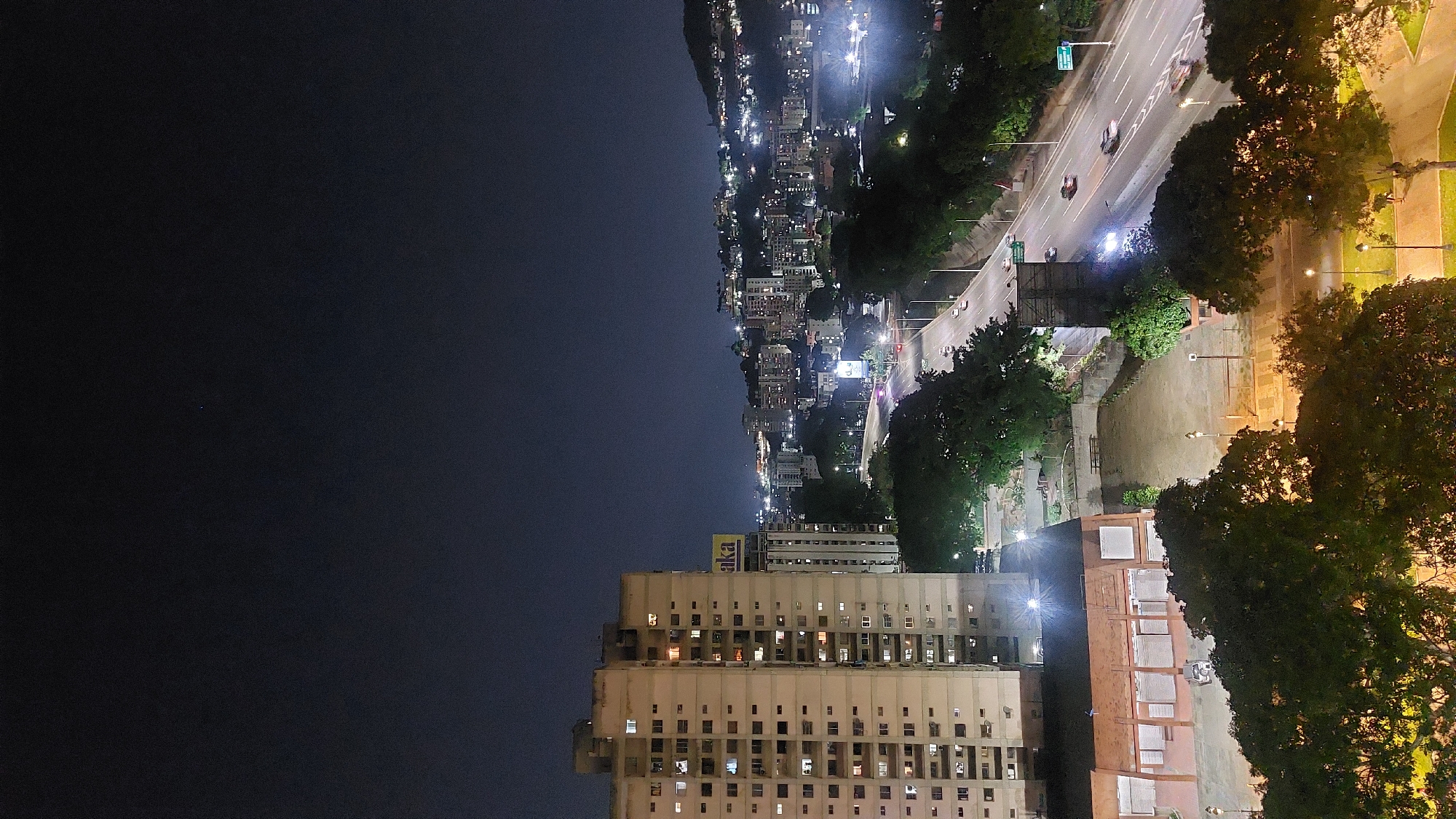 View from an elevated vantage point at night. A tall, off-white building with many illuminated windows dominates the lower portion of the frame. A highway runs parallel to the building, lined with trees and streetlights. Further away, dense urban development stretches towards the horizon, illuminated by city lights. The night sky occupies the upper portion of the image, fading into darkness. This appears to be an aerial view of Caracas, Venezuela, with the tall building possibly located in or near the city center.