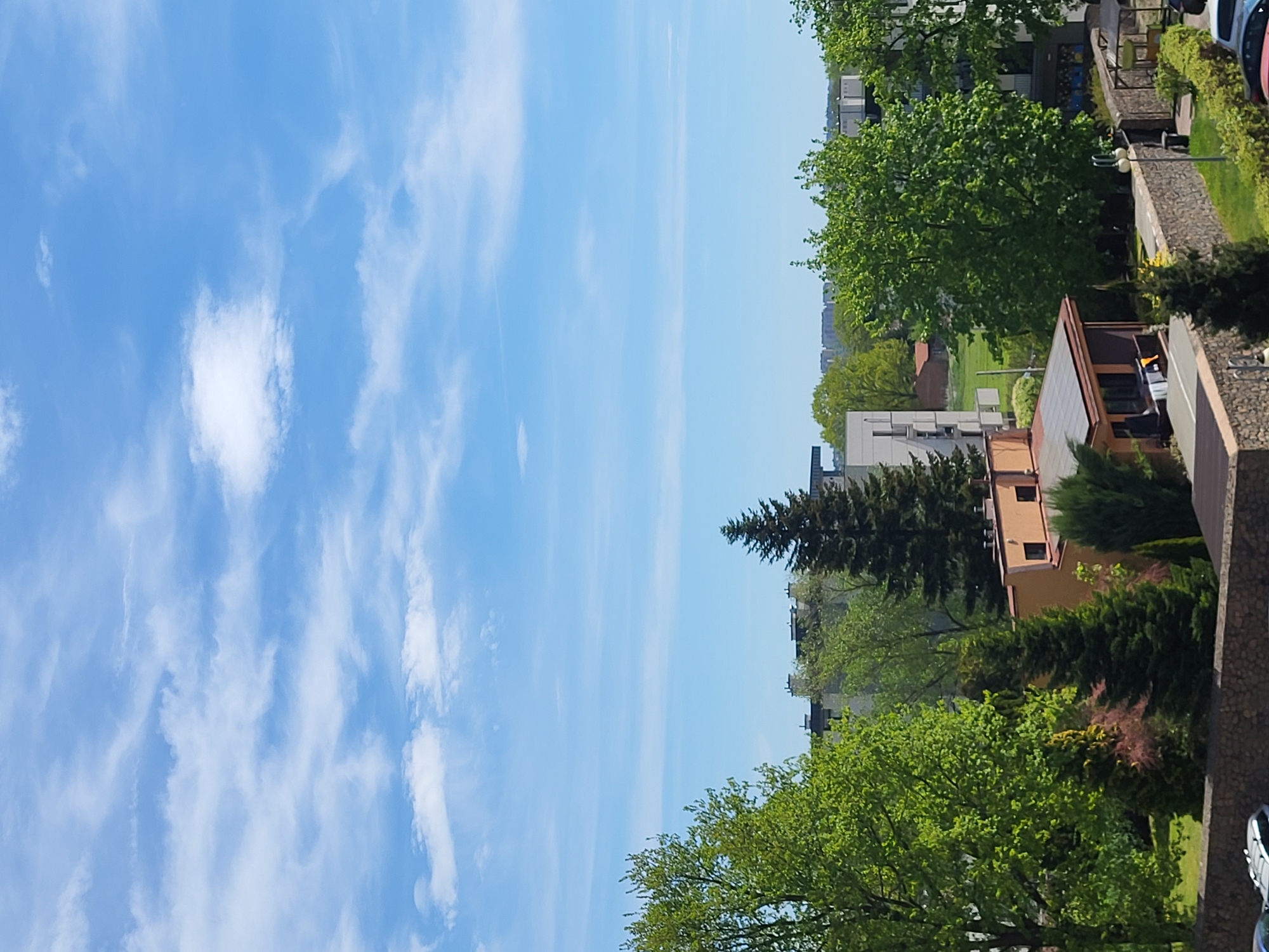 The image shows a view from a high vantage point, likely a tall building, overlooking a residential area on a sunny day. The sky occupies a significant portion of the image and is bright blue with scattered wispy clouds. Below the sky, a mix of residential buildings, trees, and greenery are visible. There are apartment buildings with flat roofs, interspersed with a variety of deciduous and evergreen trees. A brown building with visible windows is also present. In the upper right corner, portions of what appear to be balconies or rooftops with some personal items, along with cars, are visible. The location is likely a residential part of a city, but the specific city or region is not identifiable from the image alone. There are no people visible in the image.