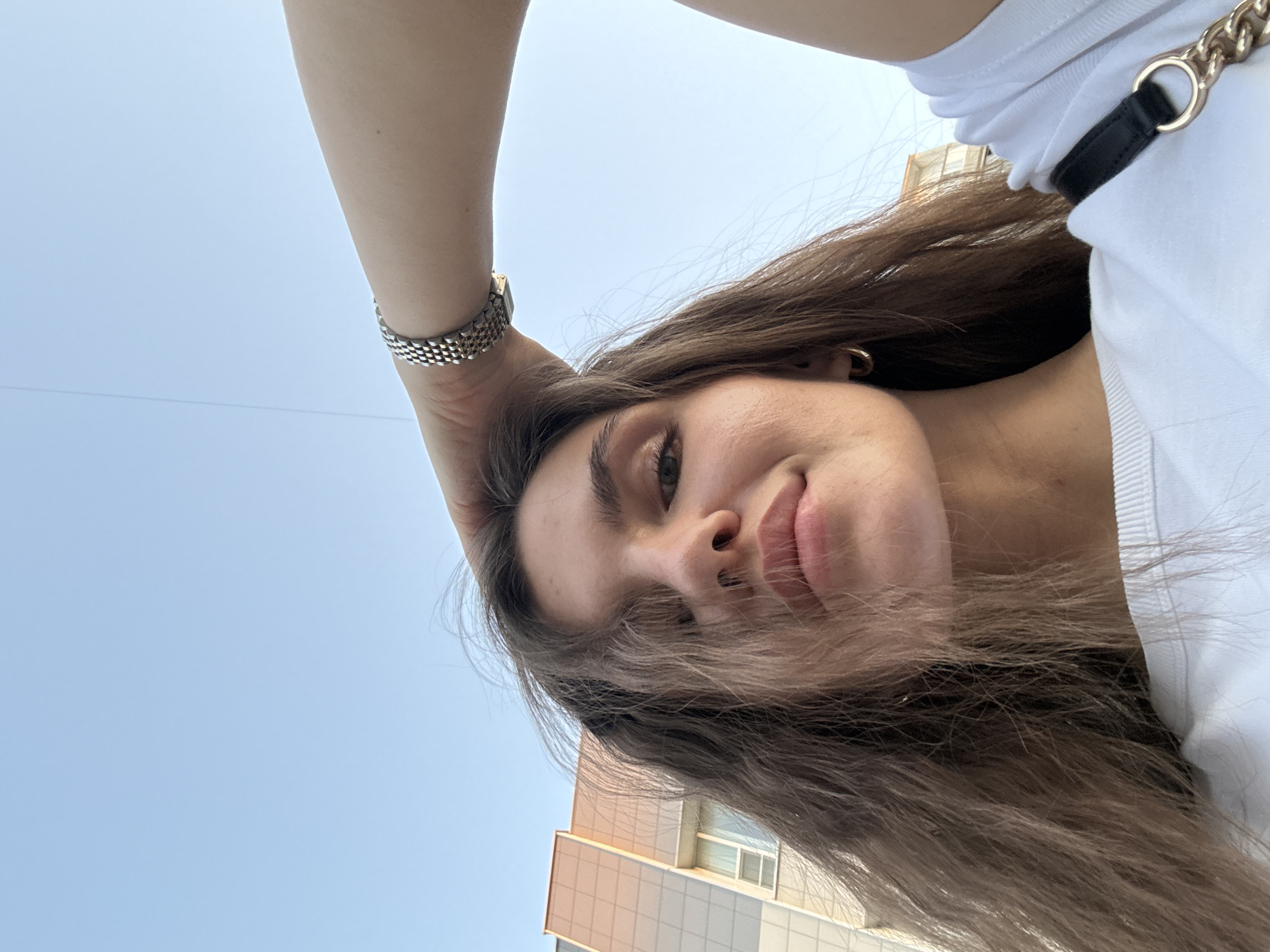 A low-angle selfie features a Caucasian woman with long, brown hair and fair skin. She is wearing a white t-shirt and a silver bracelet. Her left hand is raised to her head. A gold earring is visible in her left ear.
Behind her, a building with a grid-like facade of light brown and grey panels is partially visible. Above the building, a clear, blue sky is present. This location appears to be in an urban environment. While it's impossible to definitively confirm the location as Moscow, Russia, based on this image alone, the architectural style is consistent with buildings found in Moscow.