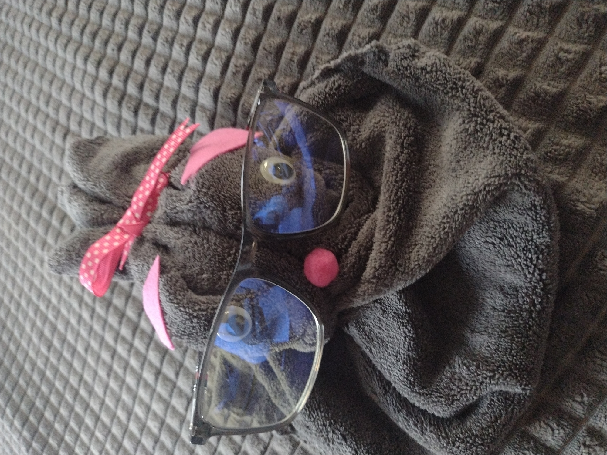 The image shows a gray stuffed animal on a gray textured surface, with a pair of glasses resting on the animal. The stuffed animal has a pink ribbon and a pink pompom on its face. The glasses reflect the room's interior. The scene is indoors. There is no indication that this image was taken in Burgas, Bulgaria.