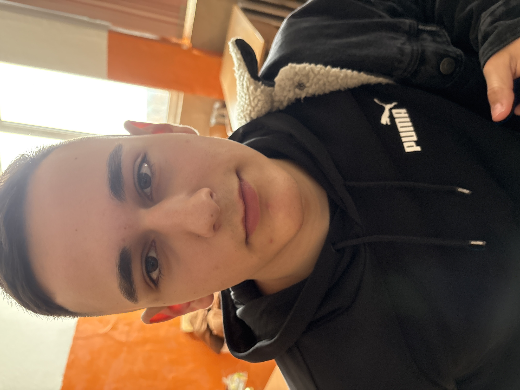 A close-up shot focuses on a young adult, facing the camera, wearing a black Puma hoodie and a black denim jacket with a light-colored sherpa collar. The background reveals a glimpse of an orange-painted wall and a window with natural light, suggesting an indoor setting. The image does not offer sufficient detail to confirm the location of Panagyurishte, Bulgaria.