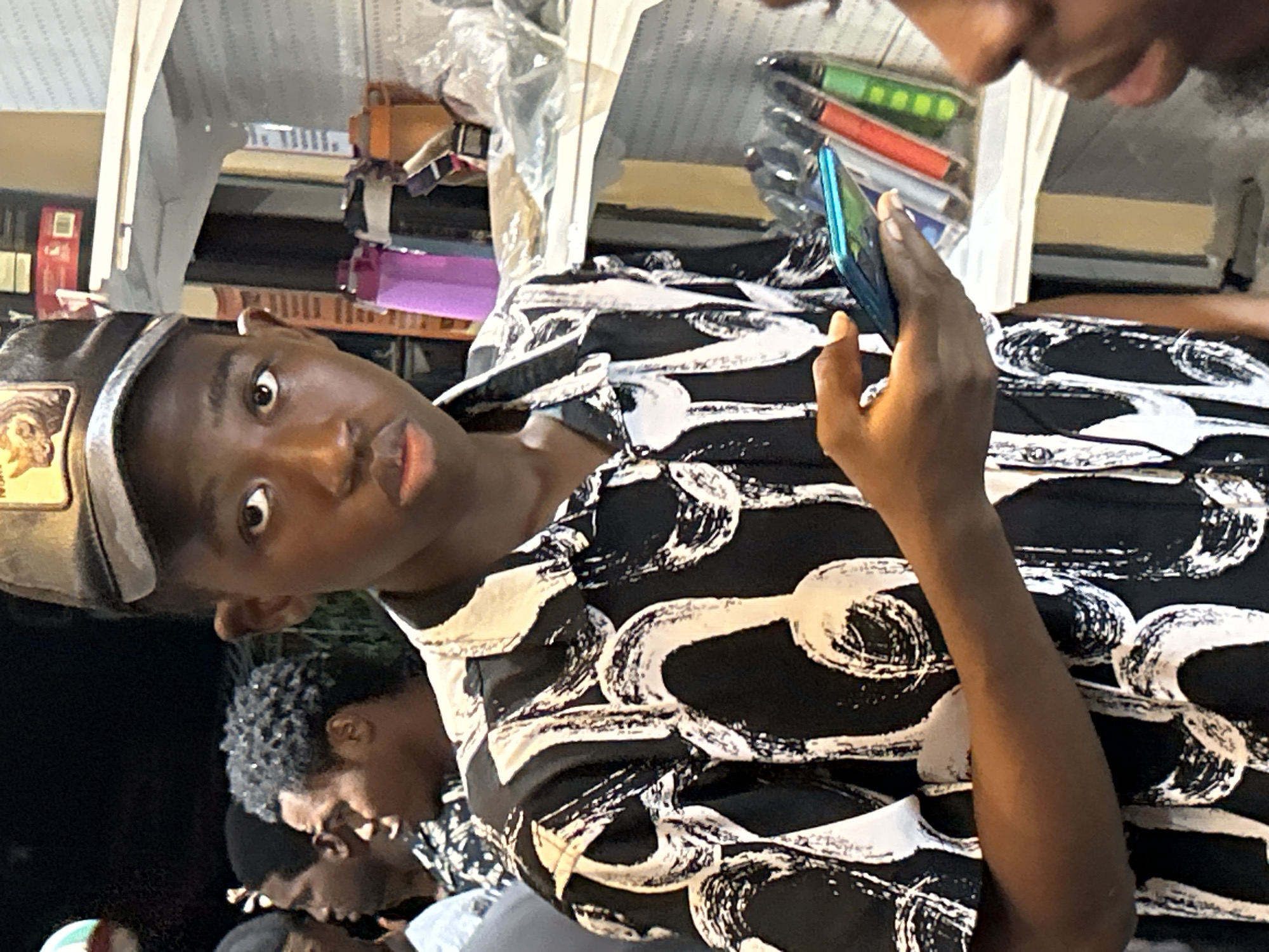 A young Black male wearing a black and white patterned shirt and a gray baseball cap is holding a smartphone in his right hand. He is looking at the camera. Behind him, to his left, are several other people. To his right, there are shelves stocked with various items, including water bottles and what appear to be vape pens or e-cigarettes. The background is dark, suggesting it may be nighttime or an indoor location with low lighting. The scene appears to be in a commercial area, possibly a shop or market stall, located in Ihiagwa, Nigeria.