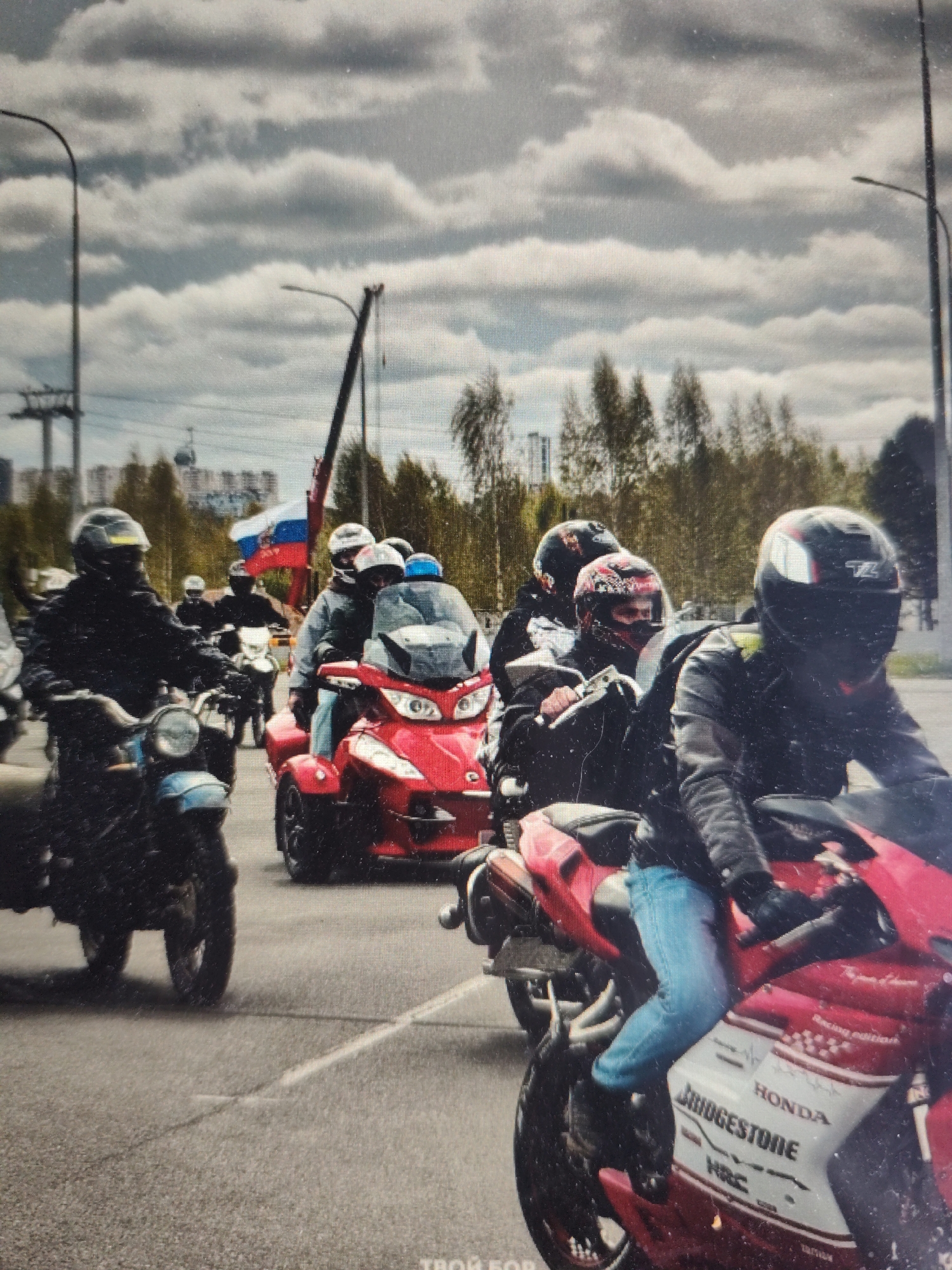 A group of motorcyclists is riding on a paved road, likely in the city of Bor, Russia, under a cloudy sky. The foreground features a red three-wheeled motorcycle with a rider wearing a black helmet and a black leather jacket with yellow accents, along with light blue jeans. The red motorcycle has the "HONDA" and "BRIDGESTONE" logos visible on its side. To the left, a rider on a black motorcycle is wearing a black helmet and a black jacket. Behind them, other motorcyclists are visible, some on motorcycles and one on a red three-wheeled vehicle. A Russian flag is also prominently displayed, attached to a pole and flying from a crane in the background. Trees and buildings are visible in the midground, and power lines and a cable car are present in the far background, suggesting an urban or semi-urban setting. The road surface appears to be asphalt with some visible white lane markings.