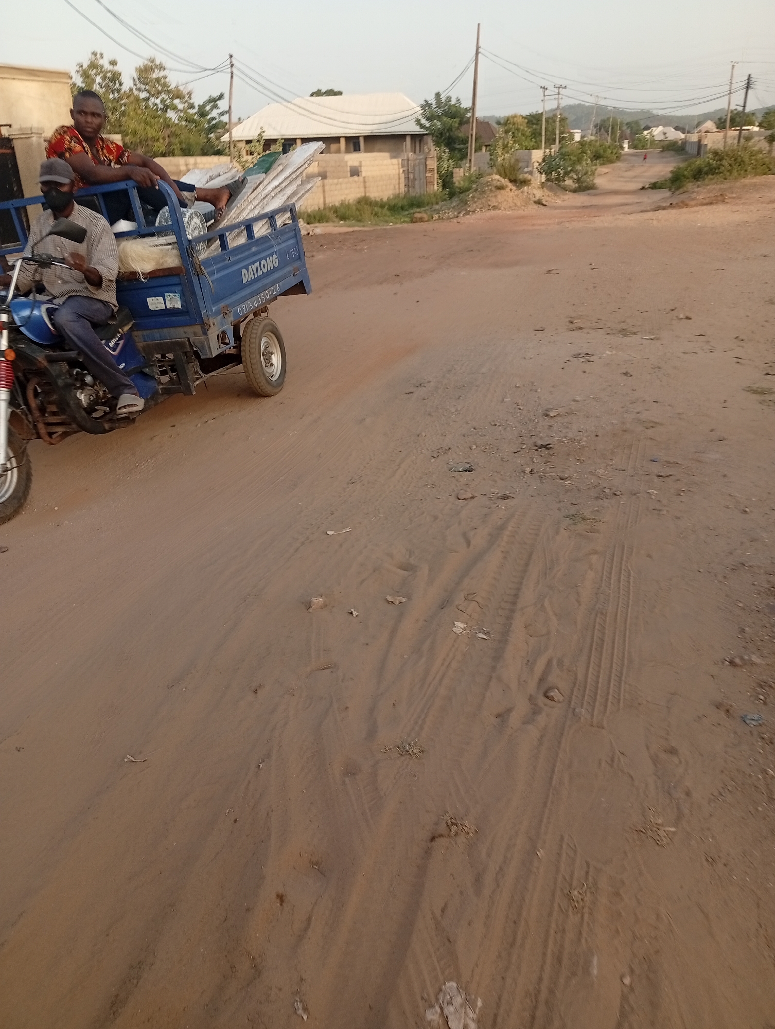 The image depicts a road in Adankolo, Nigeria, with a blue "DAYLONG" three-wheeled vehicle loaded with metal sheets and other materials on the left side of the frame. Two men are on the vehicle; one is driving, wearing a face mask and a light-colored shirt, while the other is sitting on the cargo wearing a patterned shirt. The road is sandy and dusty, showing tire tracks and scattered debris. In the background, there are buildings, trees, and utility poles. The sky is overcast.