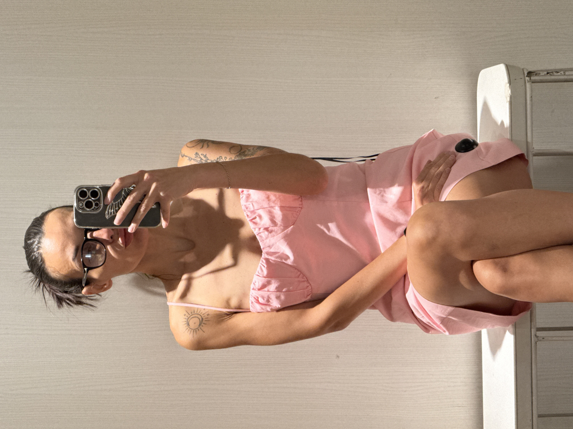A woman with dark hair pulled back, wearing glasses and a pink dress, sits on a white bench and takes a mirror selfie with her phone. She has tattoos on her left arm and shoulder. The background is a light-colored, textured wall. The scene appears to be indoors, possibly a fitting room, with a bright light source casting shadows on the wall and the woman's body. There are no discernible elements that would indicate a location in Pleven, Bulgaria.
