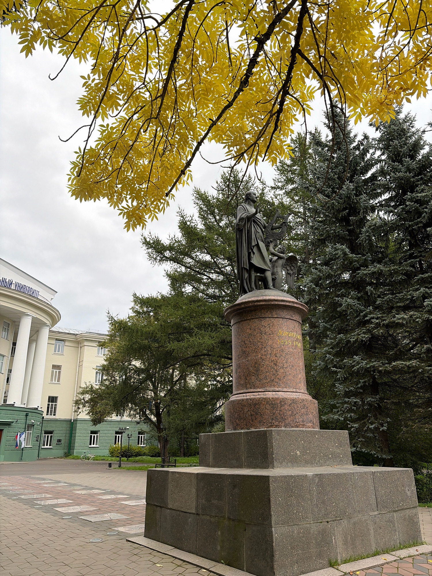 The image captures an outdoor scene featuring a statue of a man and a child in front of a building and trees. The statue, made of bronze, stands on a granite pedestal. The man is depicted holding a lyre, and the child appears to be standing next to him. The building in the background is a light yellow color with white columns and windows. It has text written on it in blue Cyrillic letters, reading "СЕВЕРНЫЙ (ФЕДЕРАЛЬНЫЙ) УНИВЕРСИТЕТ", which translates to "Northern (Federal) University". In front of the building, there is a green section with windows and a flag. To the right of the statue, there are several tall evergreen trees. Overhead, branches of a tree with yellow leaves are visible, suggesting the autumn season. The ground is paved with cobblestones and square tiles. There is no visible activity or interaction among people in the scene. The overall environment appears to be a public space, likely a campus or park area. This location is identified as the Northern (Arctic) Federal University named after M.V. Lomonosov in Arkhangelsk, Russia.