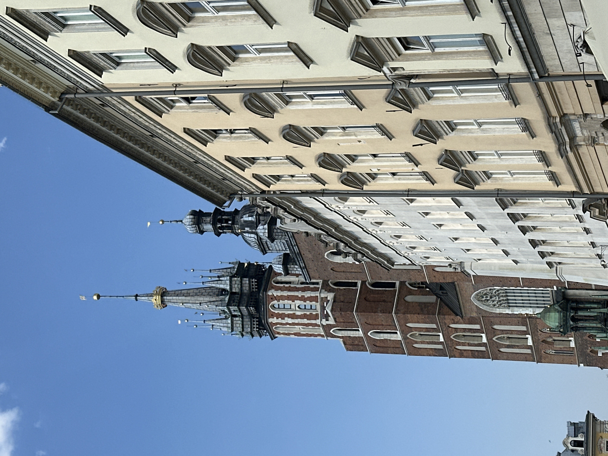 The image captures a view of St. Mary's Basilica in Krakow, Poland, seen from a low angle against a clear blue sky. The basilica's prominent brick tower with white detailing and multiple spires is on the left side of the frame. Adjacent to the basilica, on the right, are several buildings with facades in shades of yellow and cream, featuring rows of evenly spaced windows with ornate framing. The buildings exhibit architectural styles typical of European historic city centers, with decorative cornices and moldings. The foreground is dominated by the architecture, with no people or vehicles visible. The overall scene conveys a sense of historical grandeur and urban architecture.