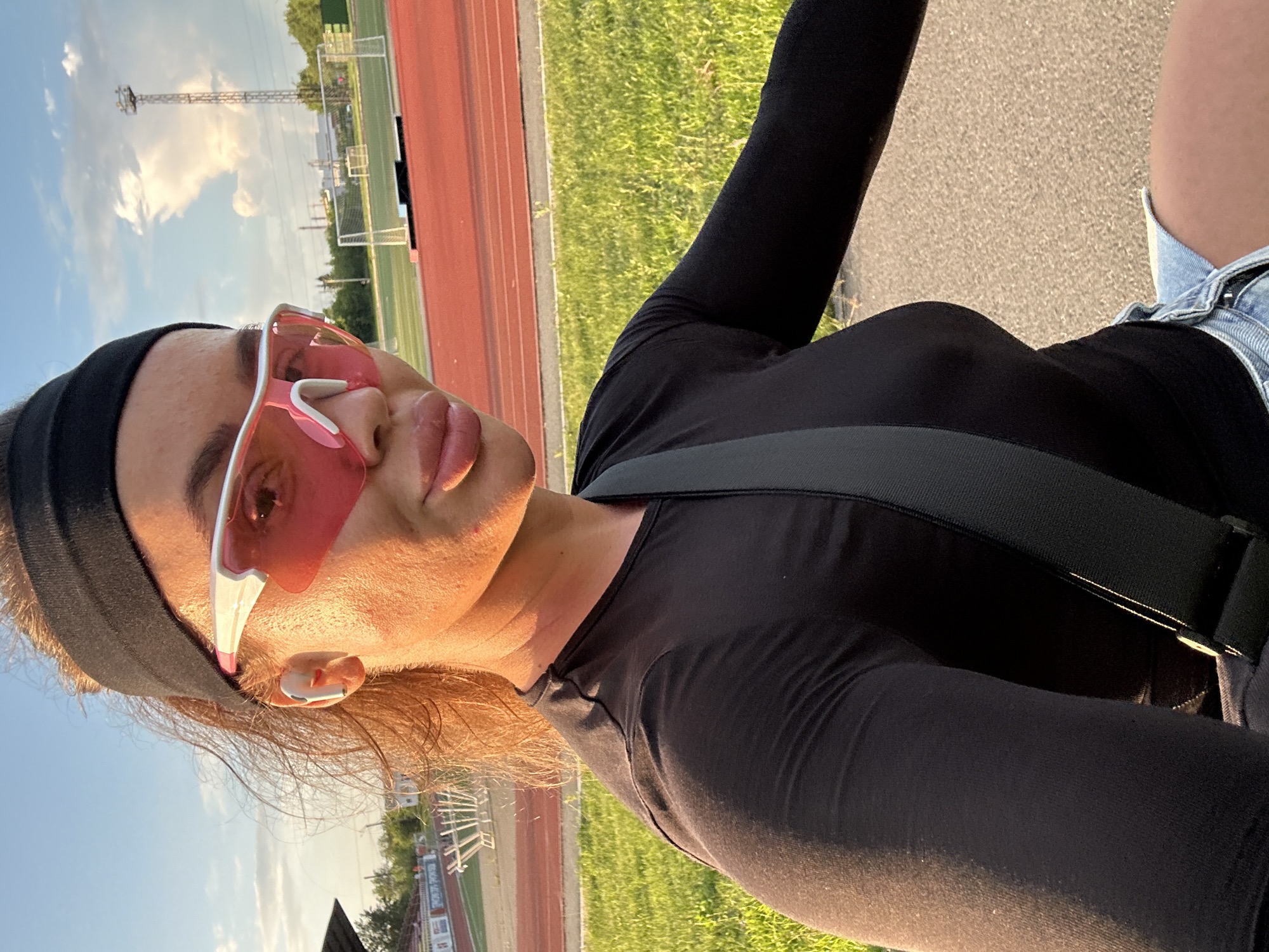 A woman with auburn hair tied back with a black headband and wearing pink sports sunglasses poses for a selfie outdoors. She is wearing a black long-sleeved shirt and denim shorts. A dark grey strap crosses her chest. Behind her, a red running track is visible, along with a grassy field, goalposts, and a tall industrial tower against a sky with scattered clouds. The scene appears to be a sports facility in Yuzhnouralsk, Russia, during the golden hour, indicated by the warm lighting.
