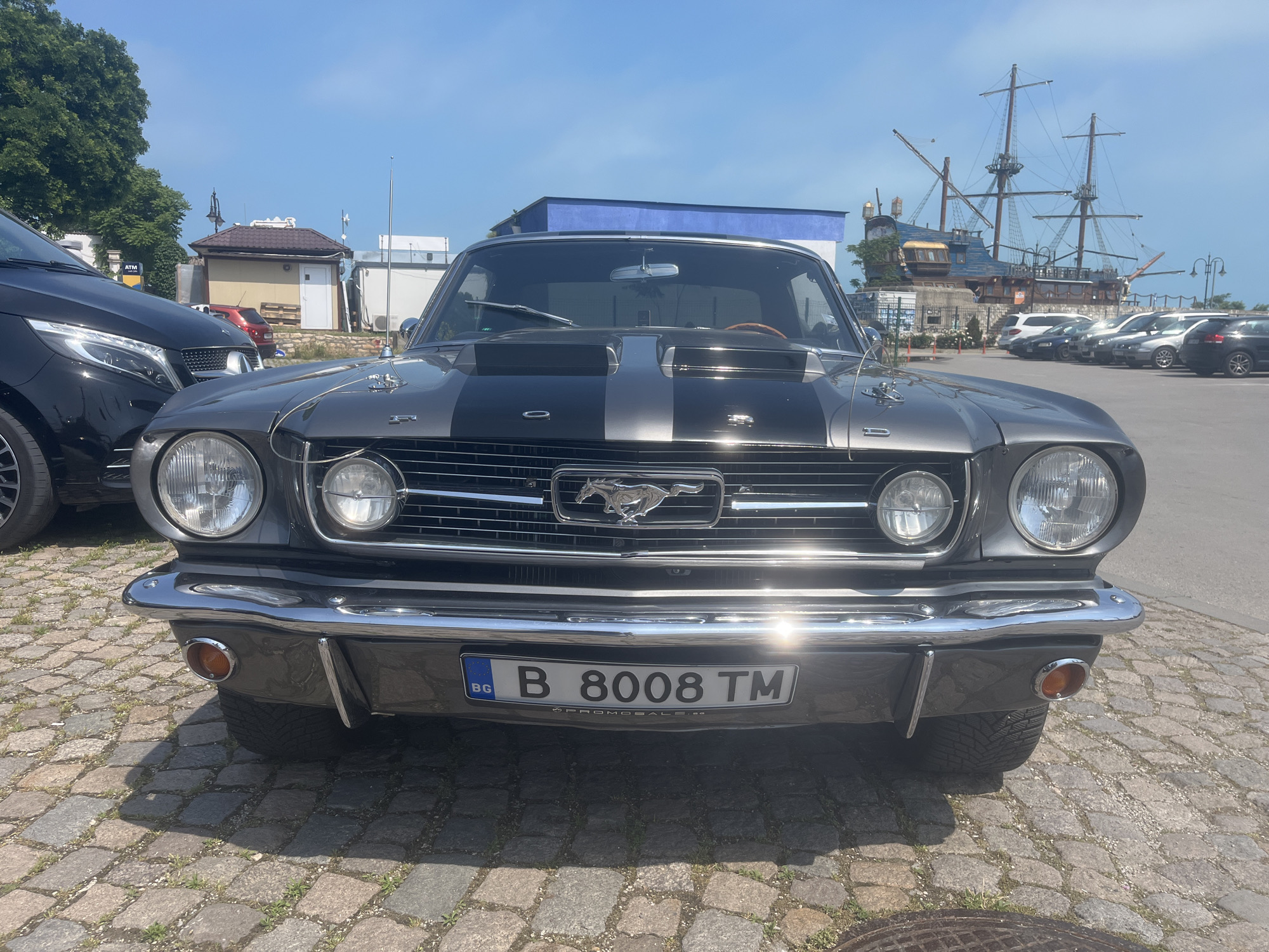 A well-maintained classic grey Ford Mustang, adorned with distinctive black racing stripes running down its hood, is prominently featured, parked head-on on a sunny day in Varna, Bulgaria. The iconic American muscle car showcases a gleaming chrome grille centered with the galloping horse emblem, complemented by chrome bumpers and round headlights. Its Bulgarian license plate, "BG B 8008 TM", is clearly visible on the front bumper, with "PROMOSALE.BG" subtly printed underneath.
The setting is a picturesque outdoor area paved with cobblestones, illuminated by bright sunlight which casts sharp shadows and highlights the car's polished surfaces. To the immediate left, the front section of a modern black Mercedes-Benz van is parked. The background is rich with details: a large, elaborate wooden structure designed to resemble a multi-masted pirate ship serves as a notable landmark, likely a restaurant or attraction. Beyond the pirate ship, several other cars are parked in a lot. Various buildings, including a small, light-colored structure with a brown roof and a building with a blue top, are scattered amidst green foliage and trees on the left, where a yellow "ATM" sign is also visible. The sky is a clear, vibrant blue, indicating fair weather. The scene captures a blend of classic automotive beauty against a lively coastal or harbor backdrop.
